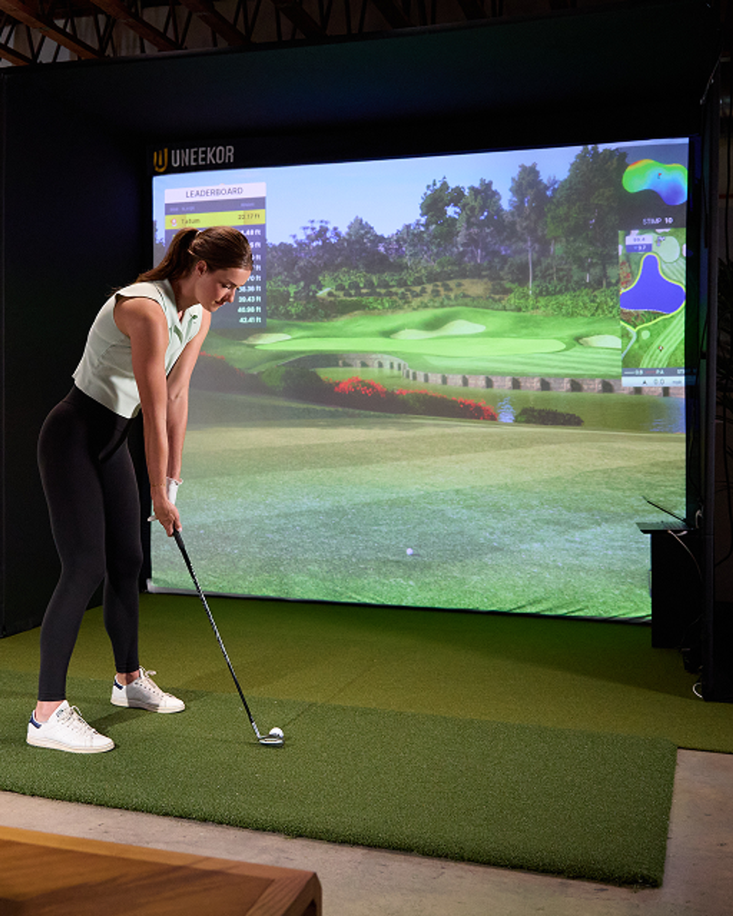 A woman addresses a golf ball on an indoor Uneekor golf simulator, displaying a virtual course on its large screen.