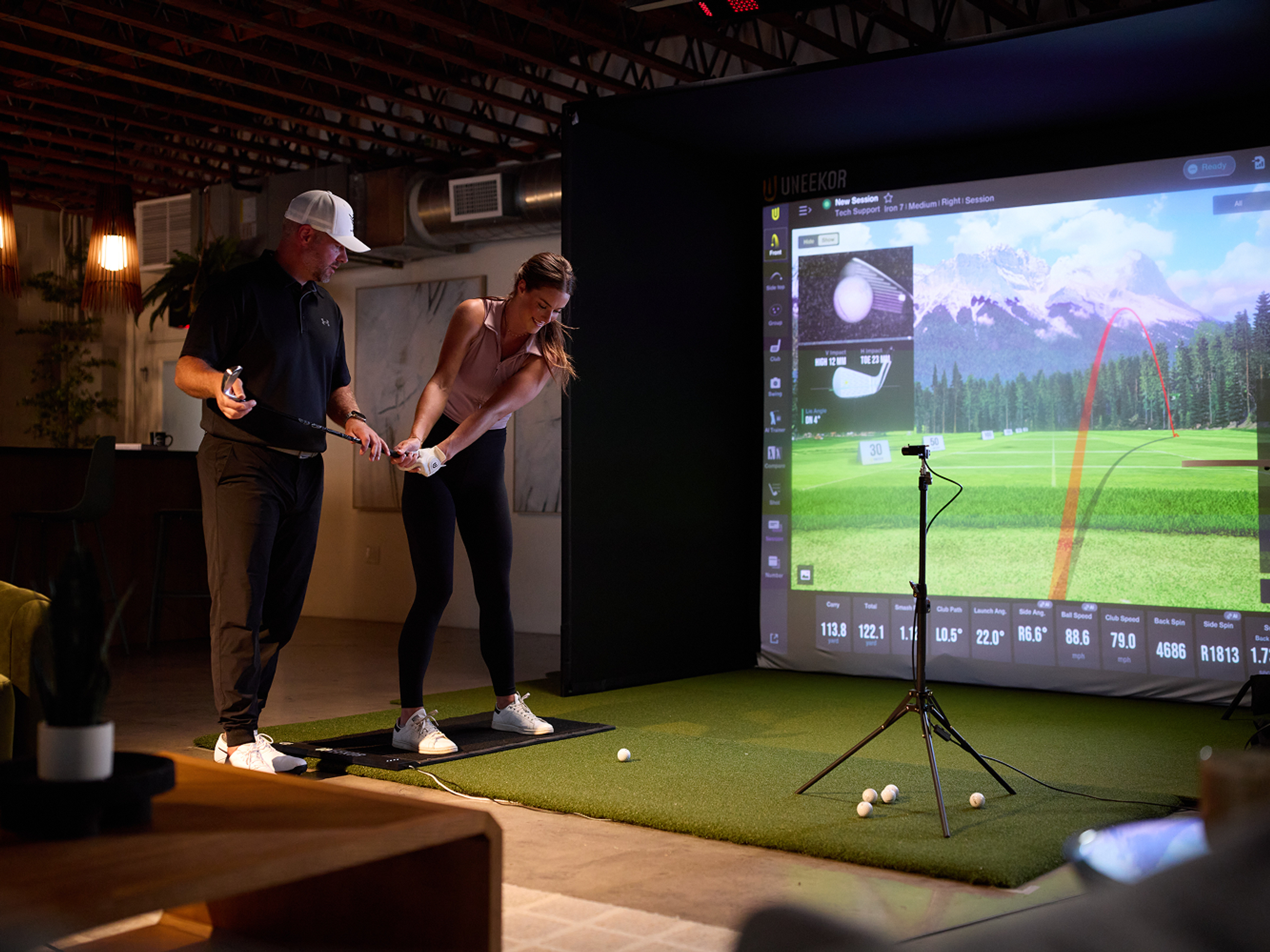 A golf instructor helps a woman practice her swing in front of a golf simulator screen displaying a virtual course.