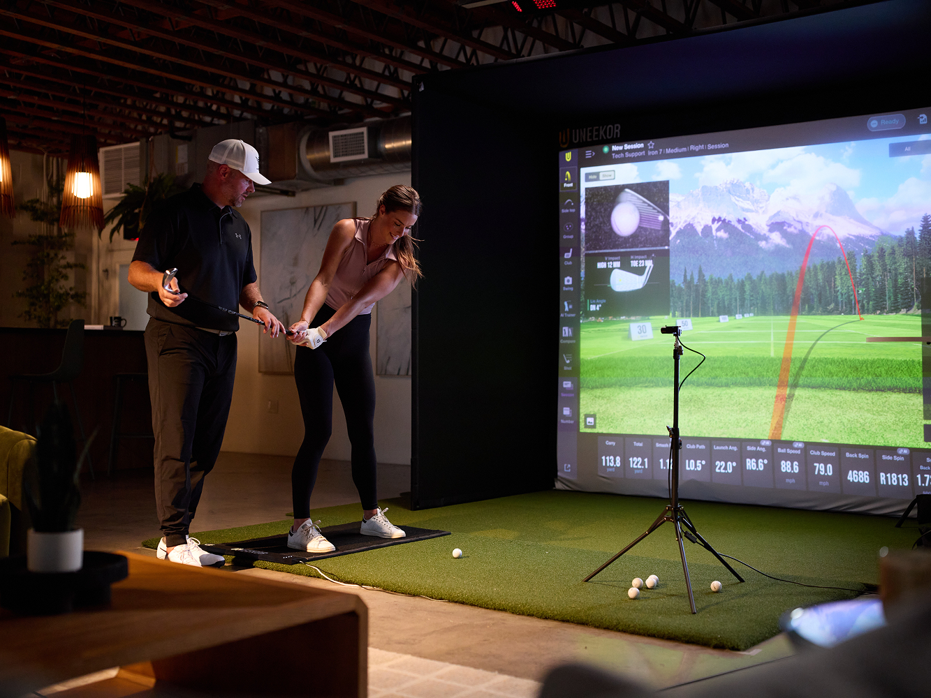 A golf instructor helps a woman practice her swing in front of a golf simulator screen displaying a virtual course.