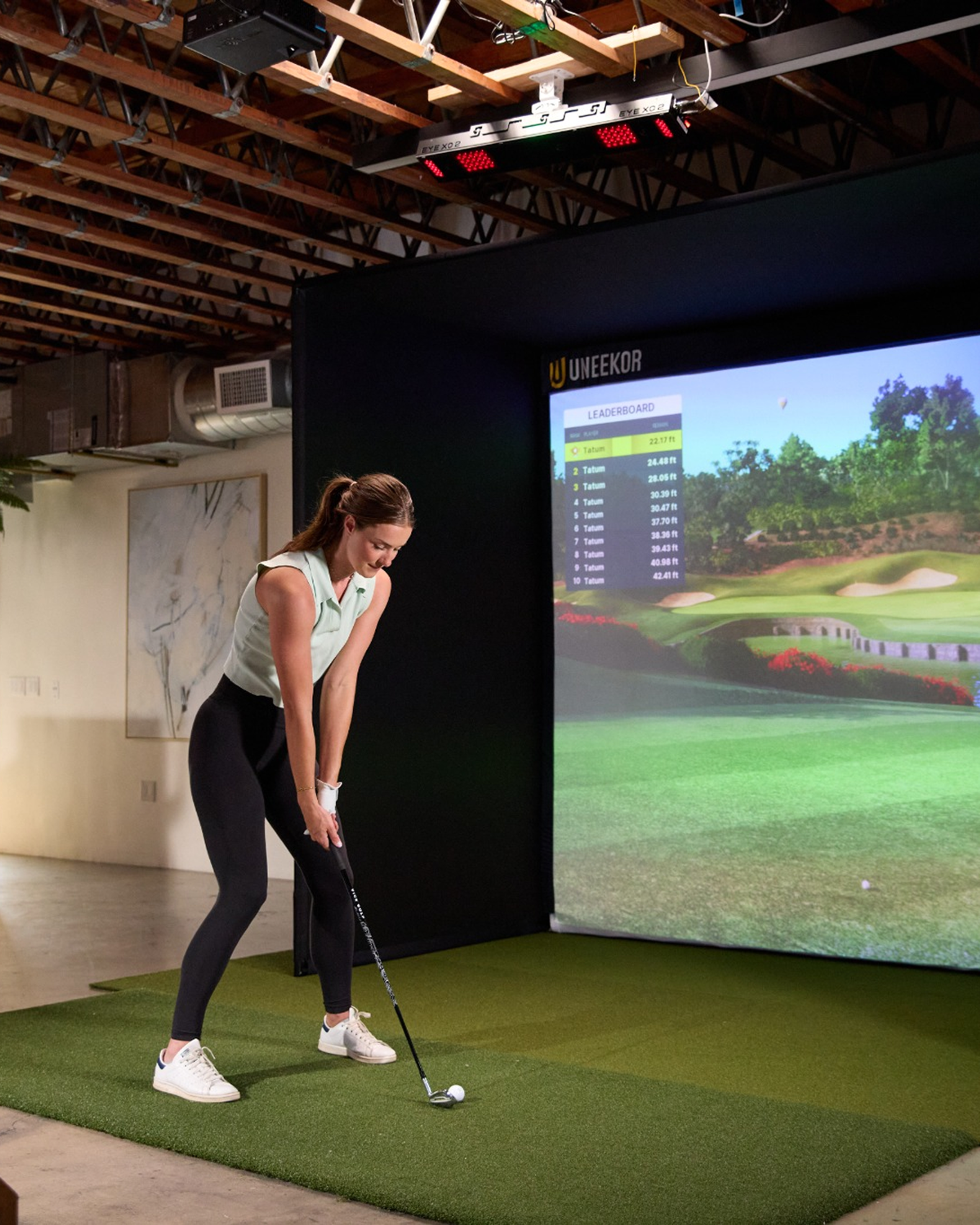 A woman plays golf on a simulator with a large screen displaying a golf course.