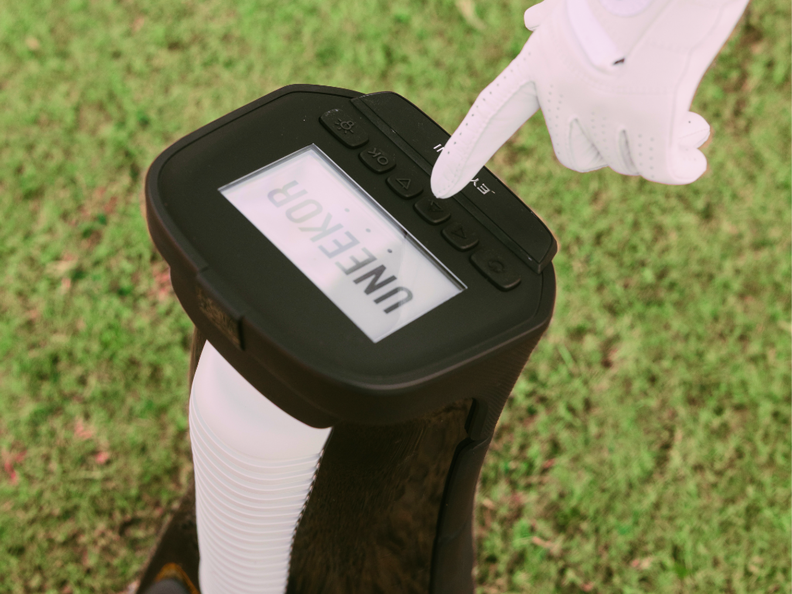 A hand in a white golf glove presses a button on a black device displaying "UNEEKOR".