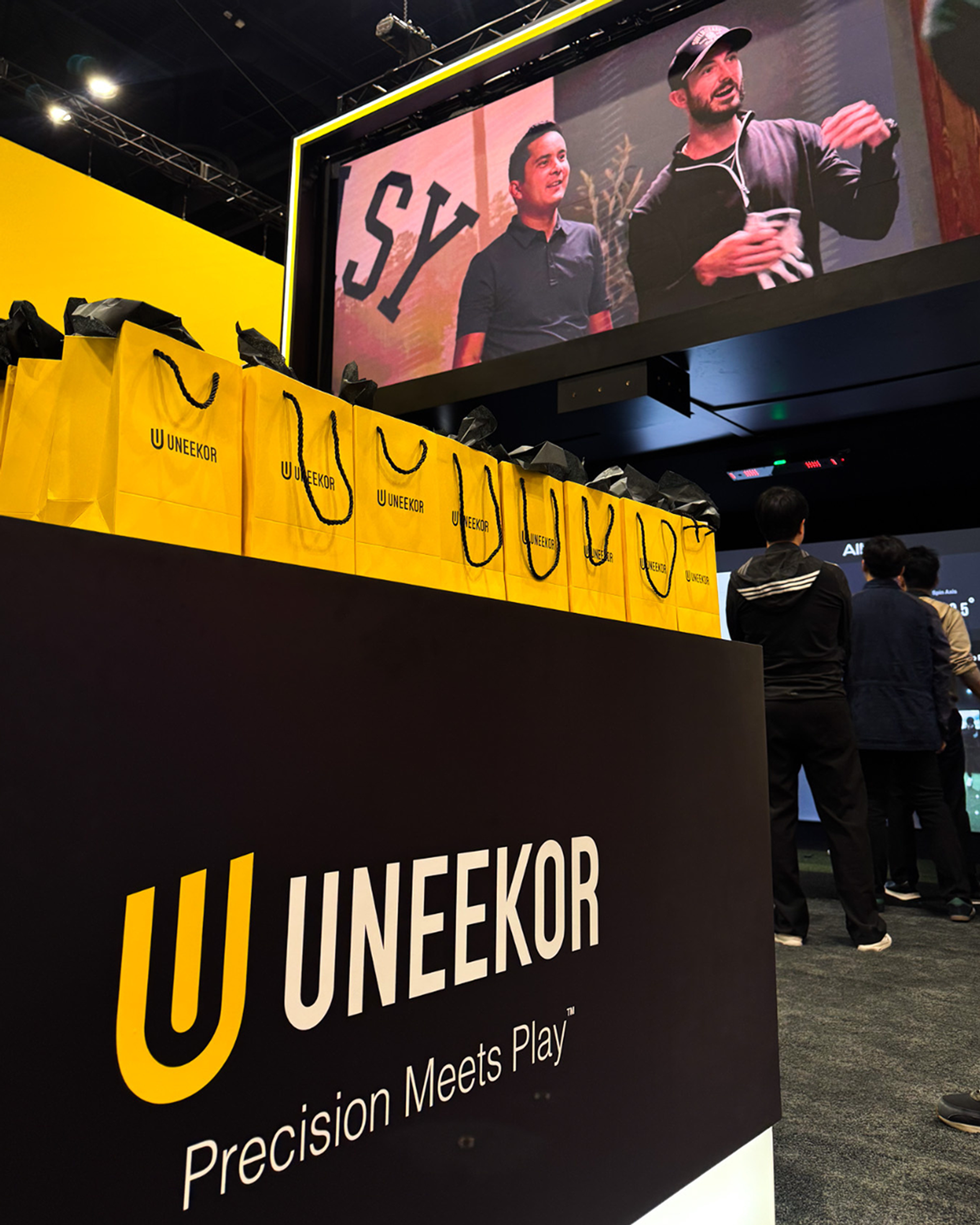 Uneekor branded display with yellow bags, a screen showing two men, and 'Precision Meets Play' slogan.