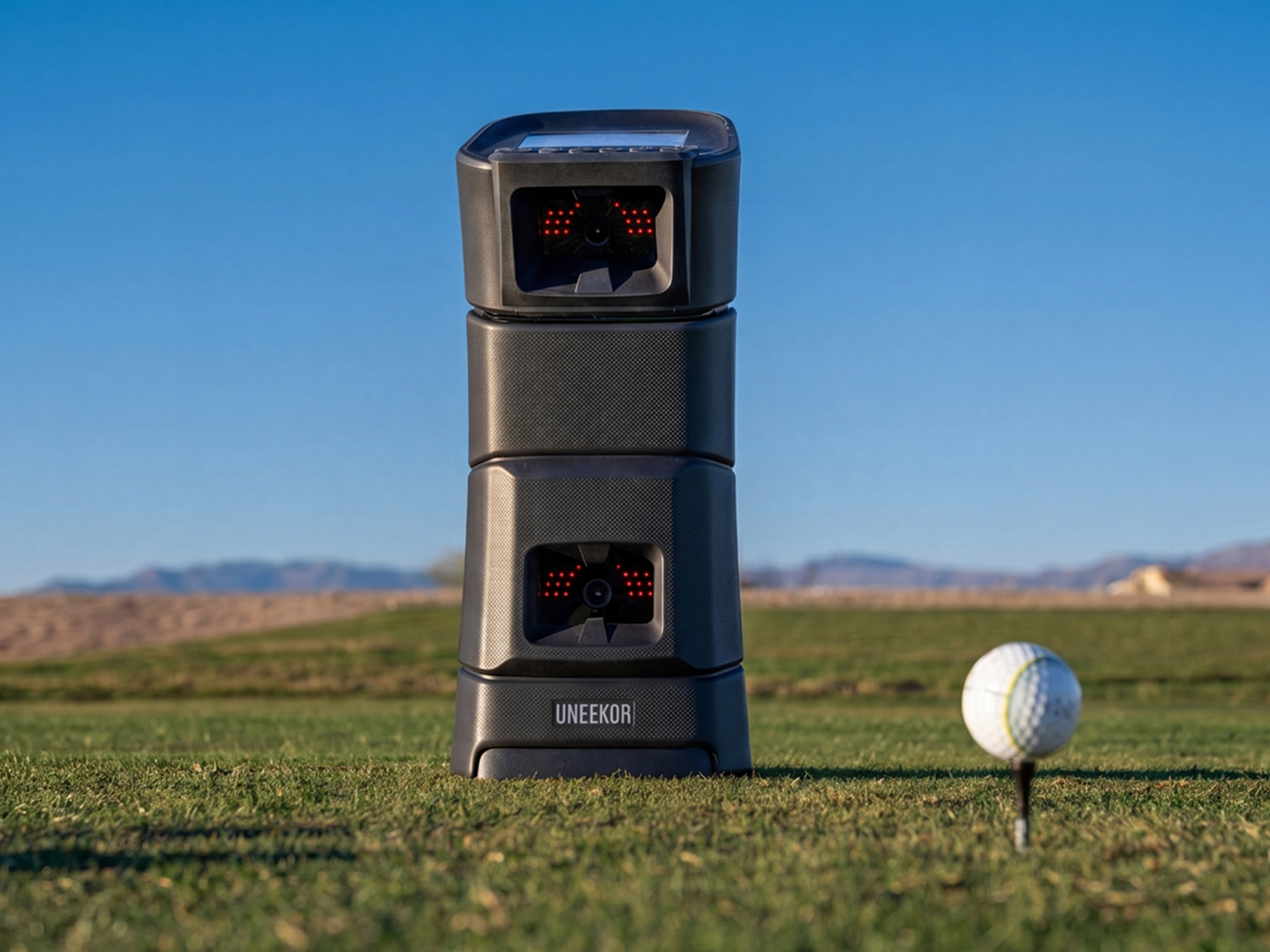A dark gray UNEEKOR golf launch monitor stands on a golf course next to a golf ball on a tee under a clear blue sky.