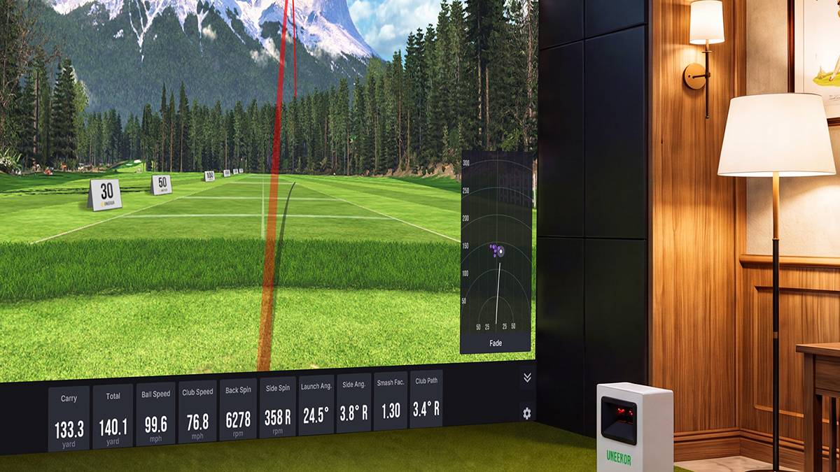 An indoor golf simulator setup with a large screen displaying a virtual golf course with mountains and a shot trajectory, a golf ball on a green mat, and a launch monitor.
