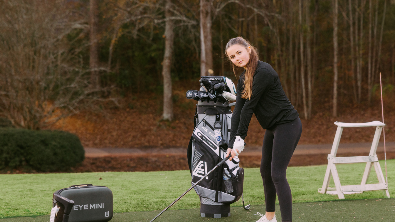 A woman in athletic wear addresses a golf ball with a driver on green turf, with an "EYE MINI" launch monitor and golf bag nearby.