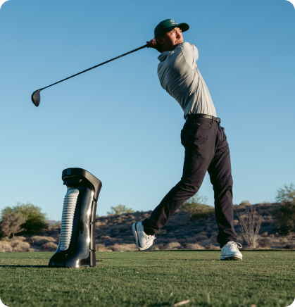 A golfer mid-swing with a driver next to a golf ball dispenser on a sunny course.