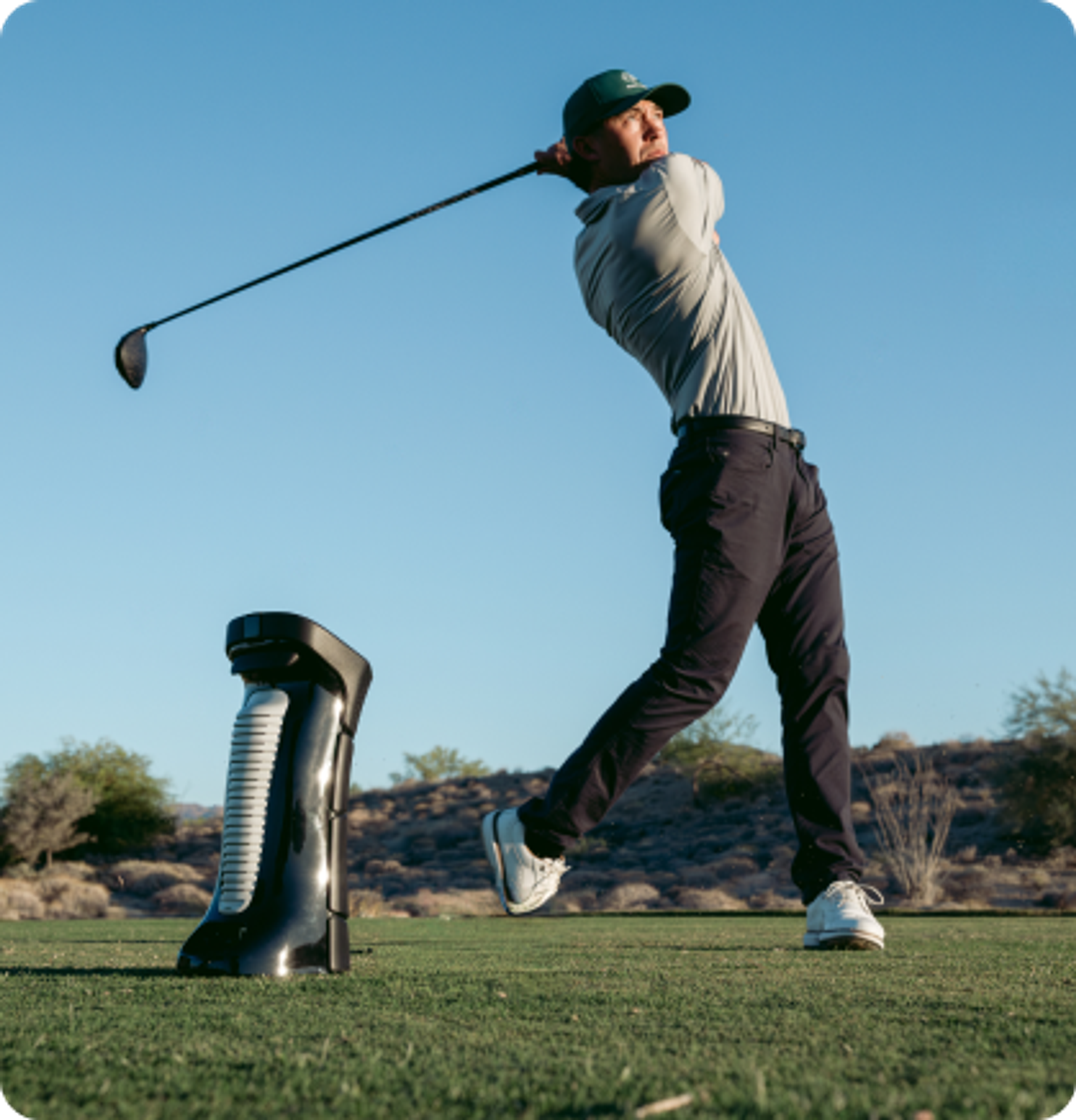 A golfer mid-swing with a driver next to a golf ball dispenser on a sunny course.