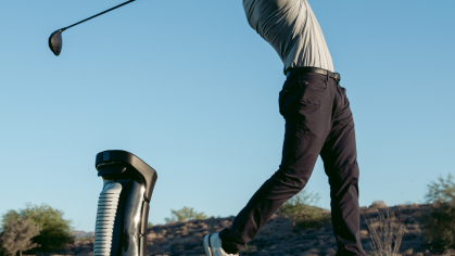 A golfer mid-swing with a driver next to a golf ball dispenser on a sunny course.