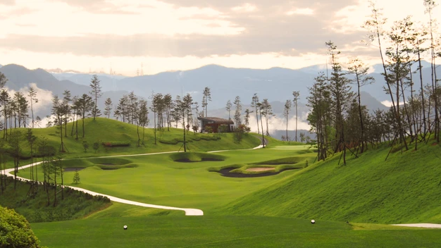Lush green golf course with a tee box, rolling hills, tall trees, and misty mountains in the background.