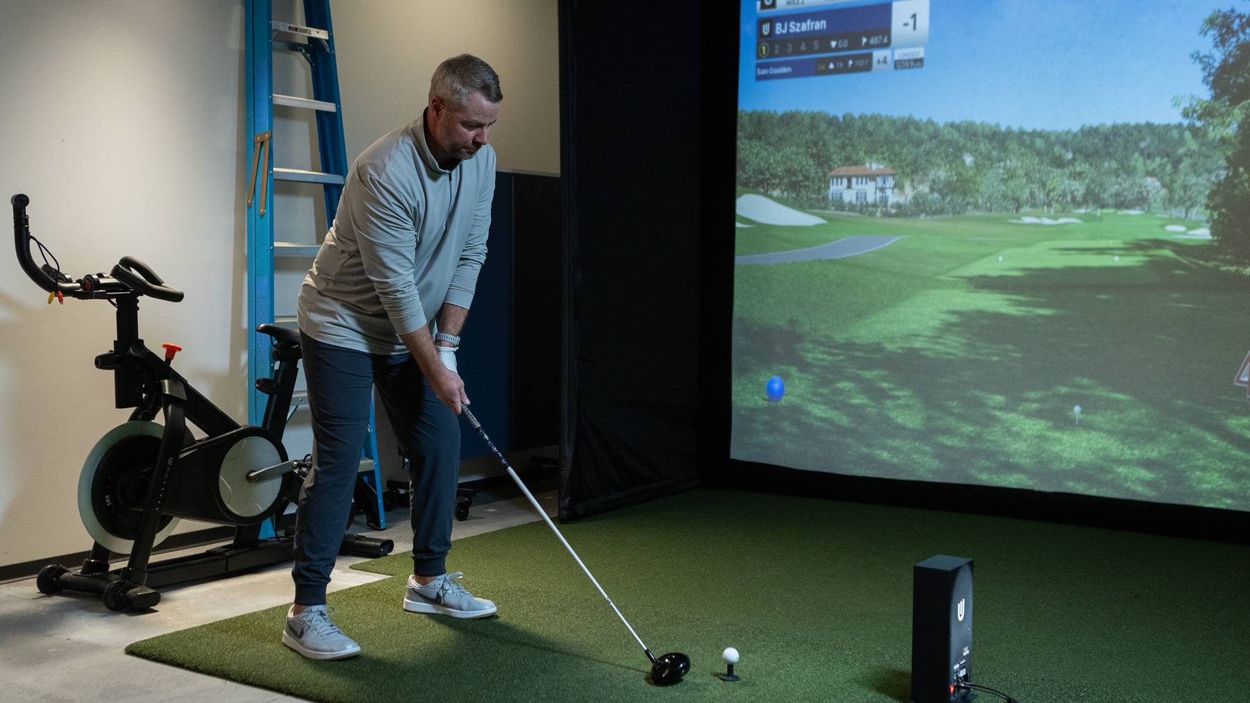 A man holds a golf club, ready to hit a ball in an indoor golf simulator showing a virtual golf course.