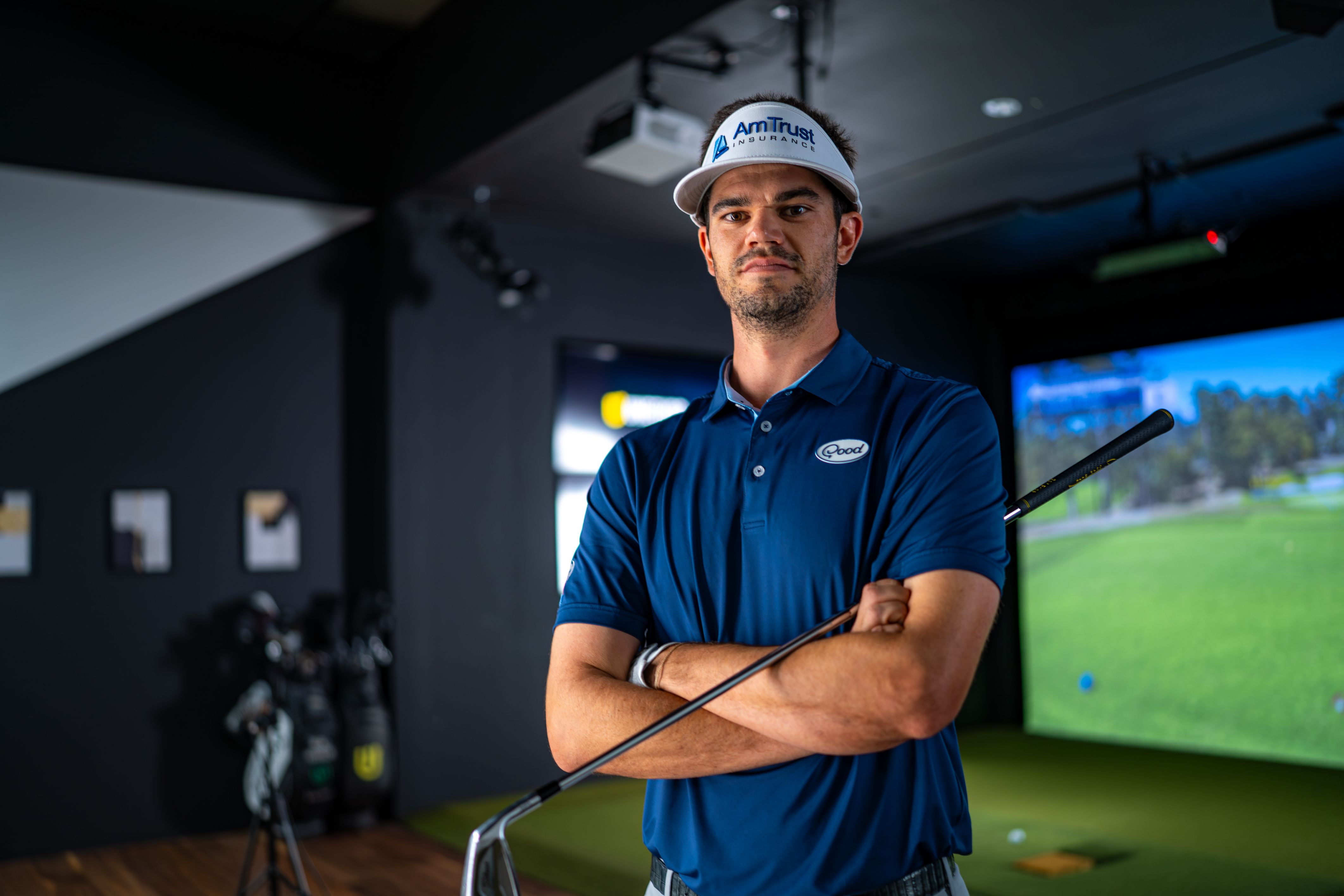 A male golfer in a blue polo and AmTrust visor stands with crossed arms, holding a golf club, in a simulator room.