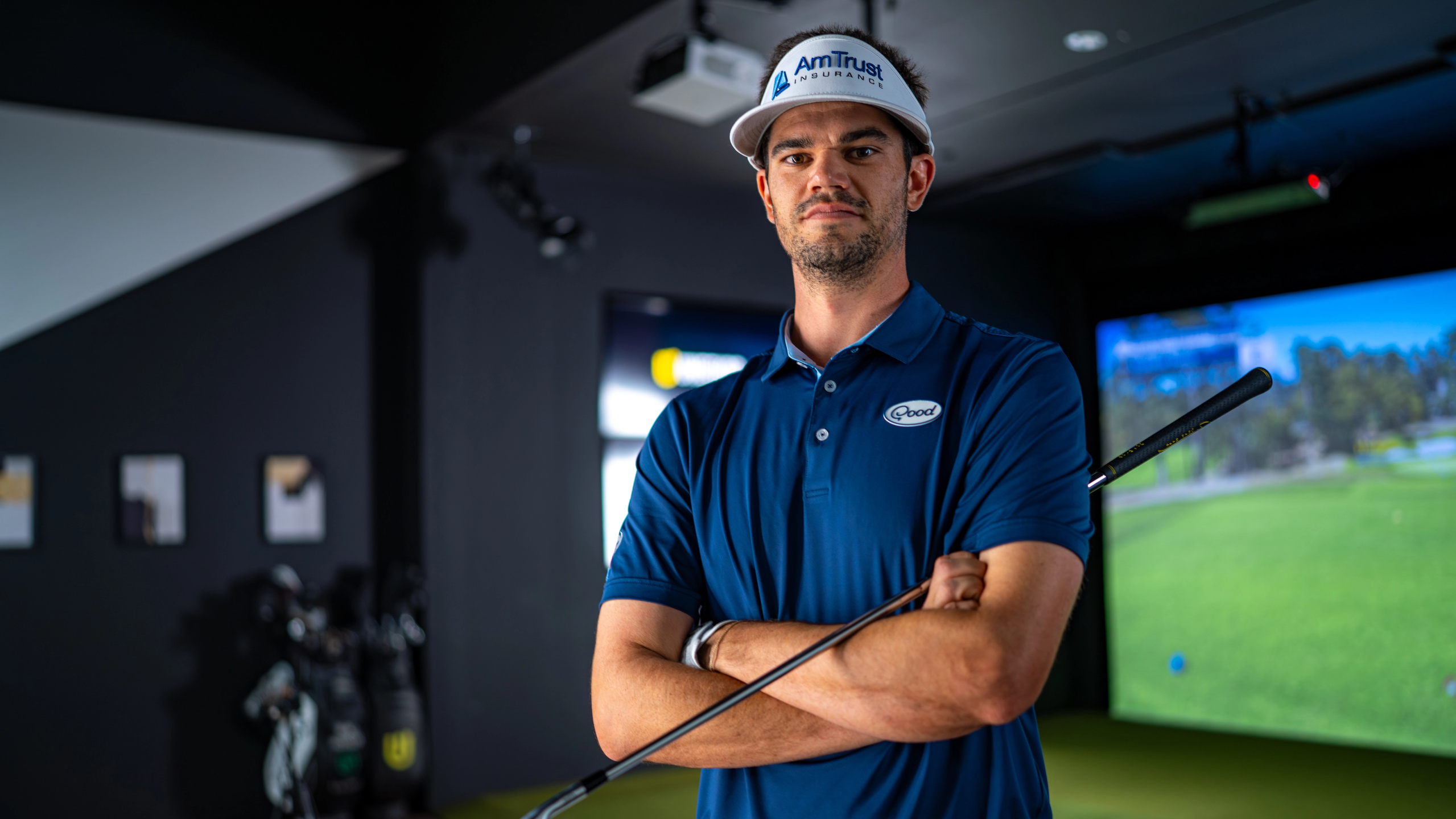 A male golfer in a blue polo and AmTrust visor stands with crossed arms, holding a golf club, in a simulator room.
