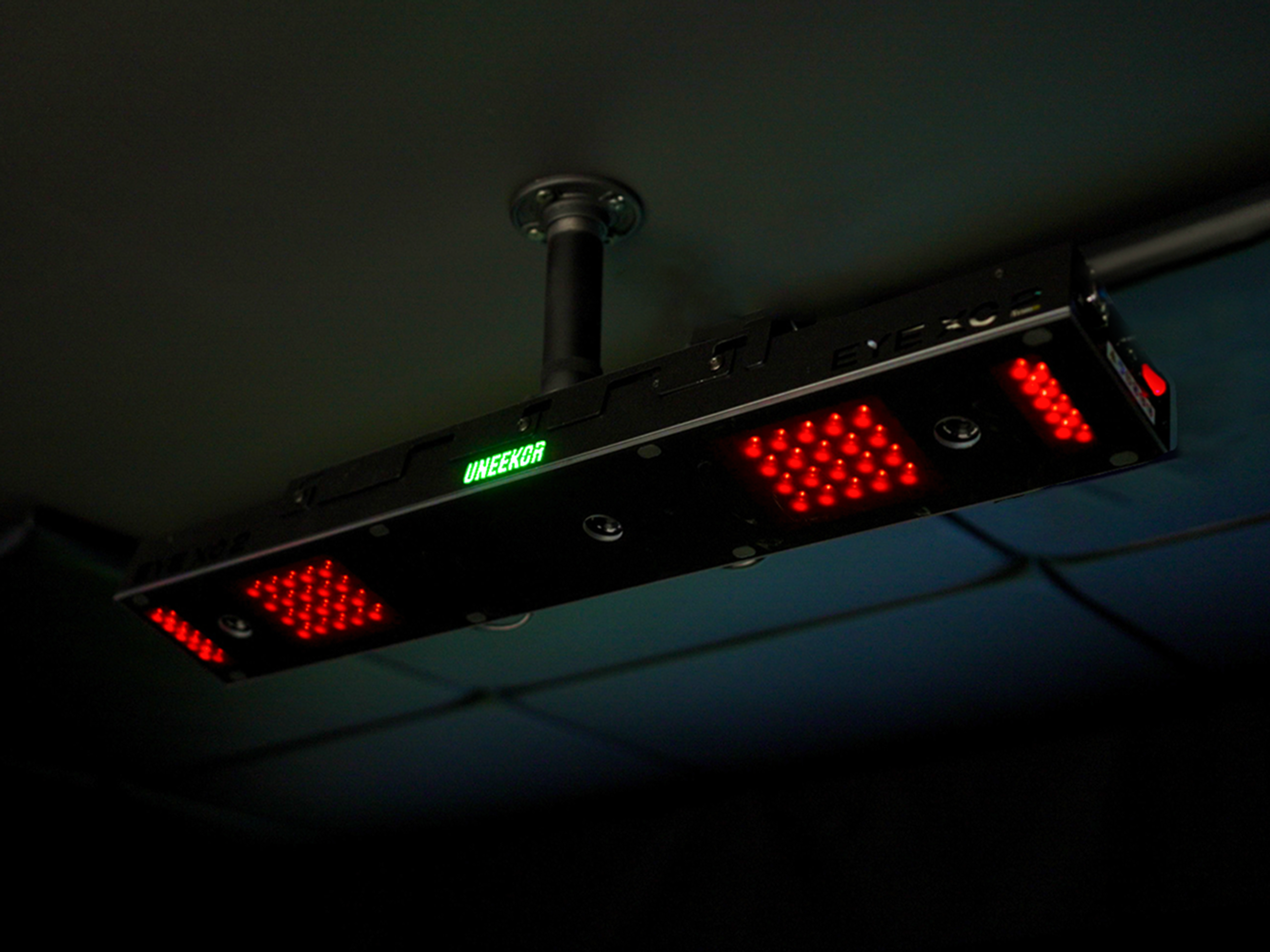 A black rectangular device with glowing red LED lights and a green 'UNEEKOR' display hangs from a dark ceiling.