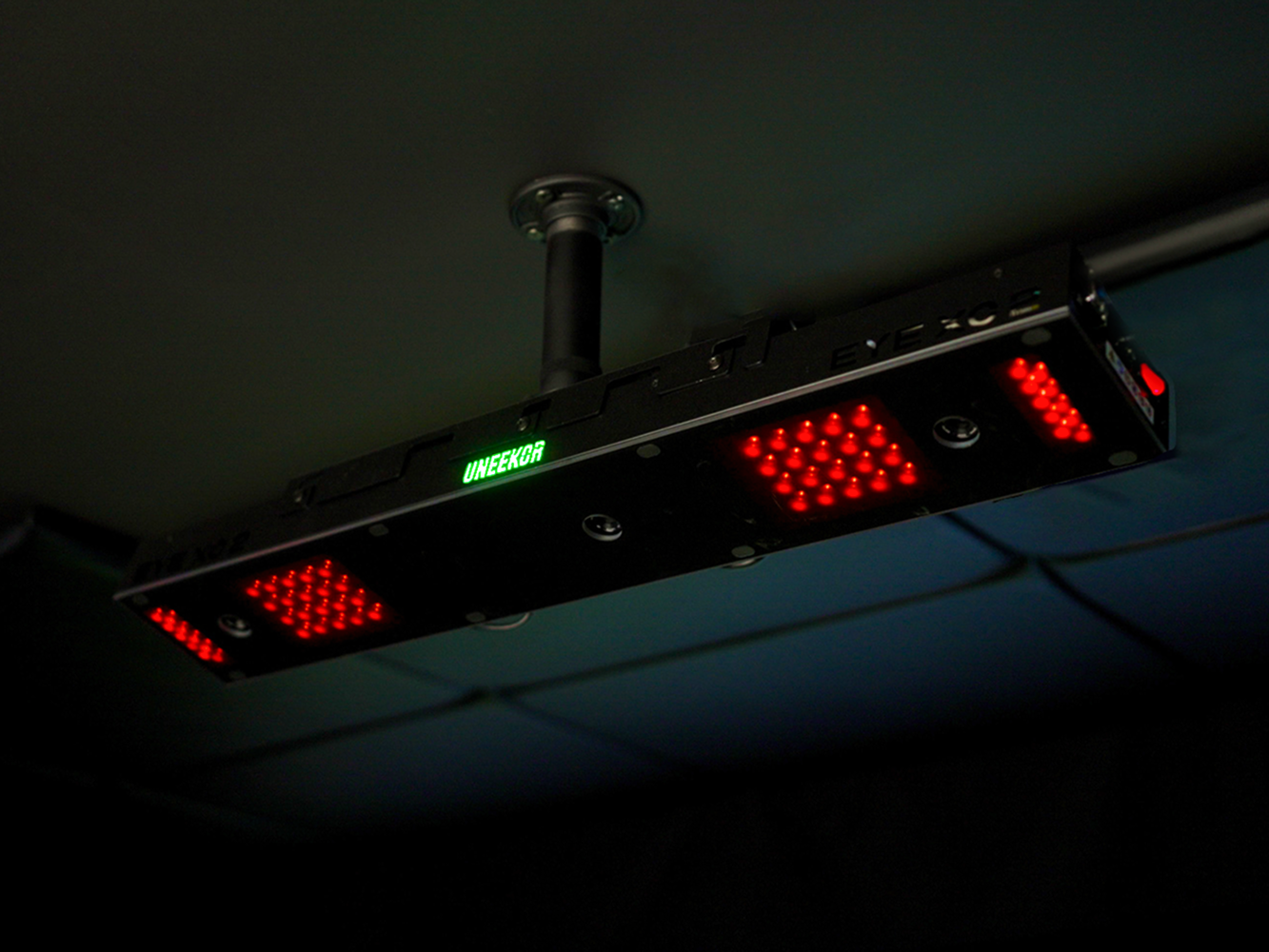 A black rectangular device with glowing red LED lights and a green 'UNEEKOR' display hangs from a dark ceiling.