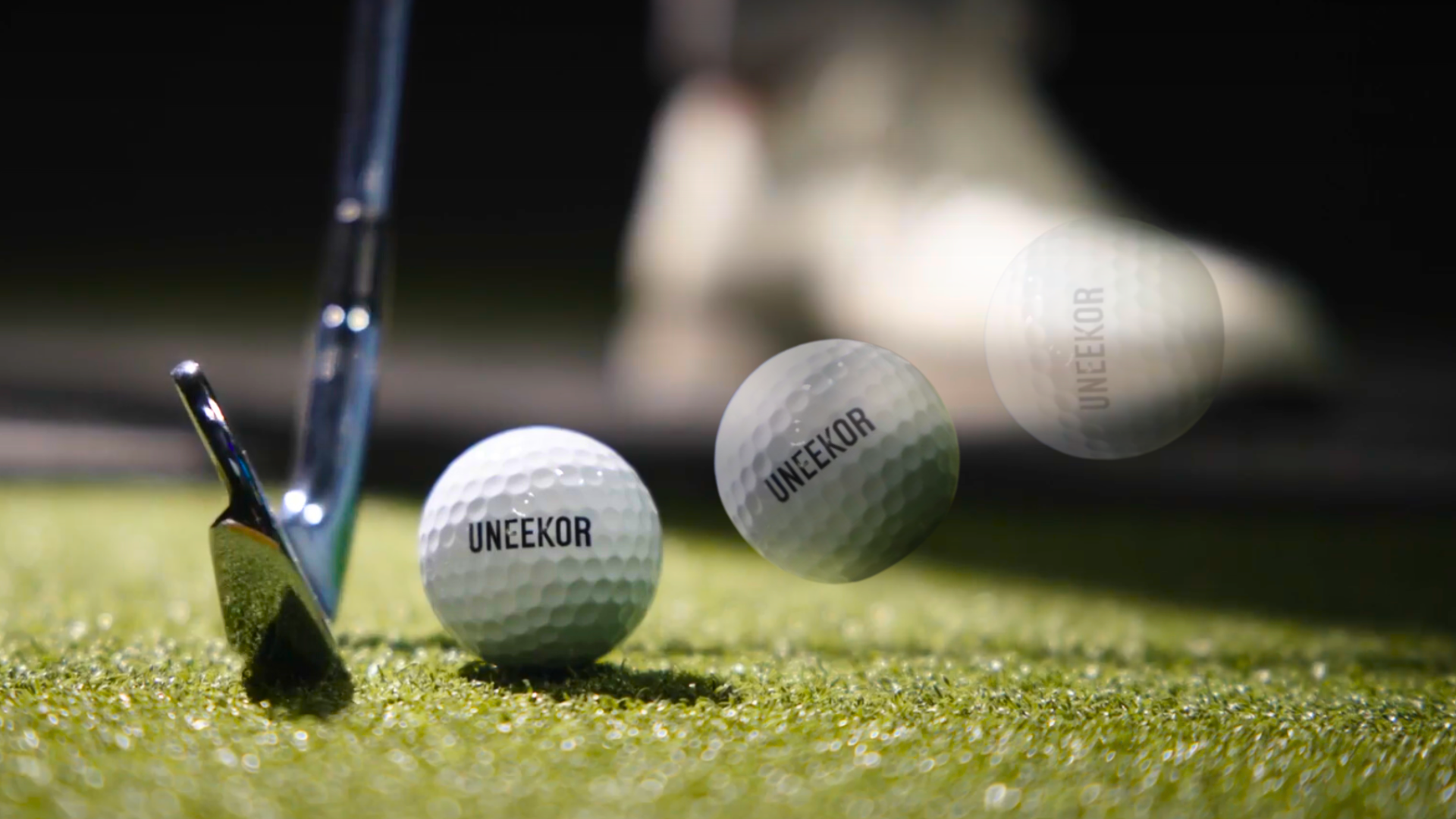 A golf club on green turf next to a golf ball, with another UNEEKOR golf ball blurred in motion.