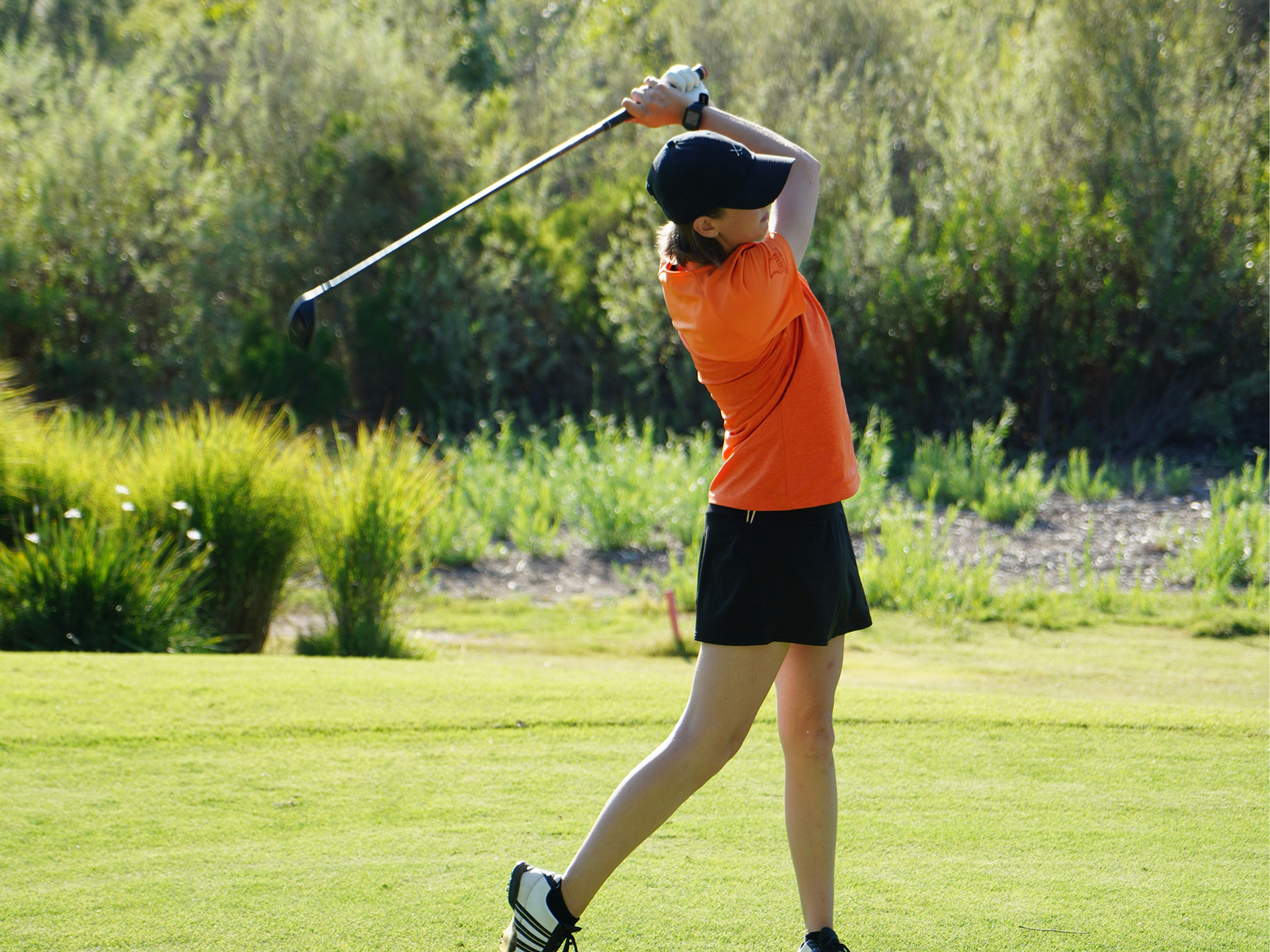 A female golfer in an orange shirt mid-swing on a green golf course.