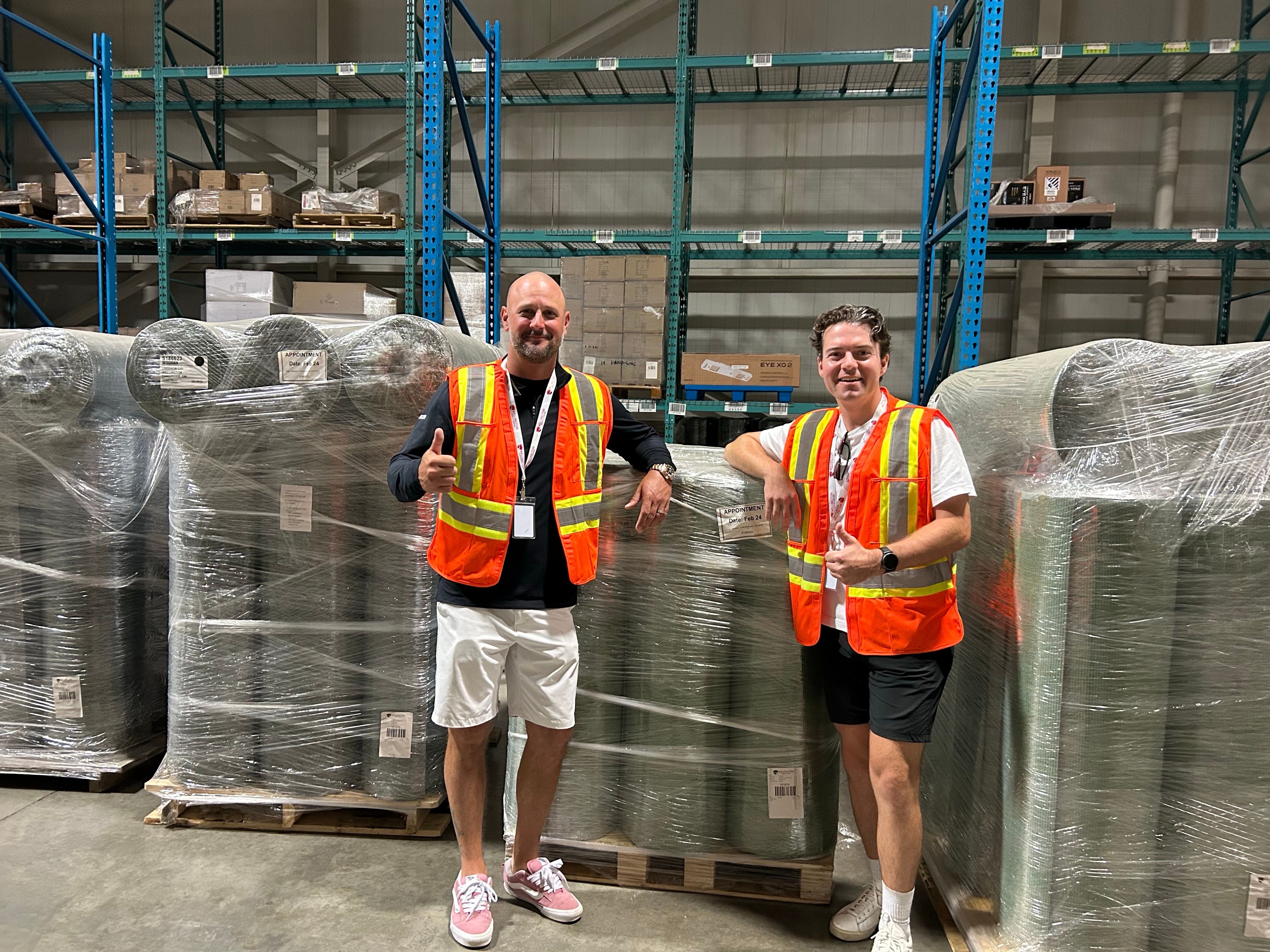 Two men in orange safety vests smile in a warehouse next to pallets of rolled materials.