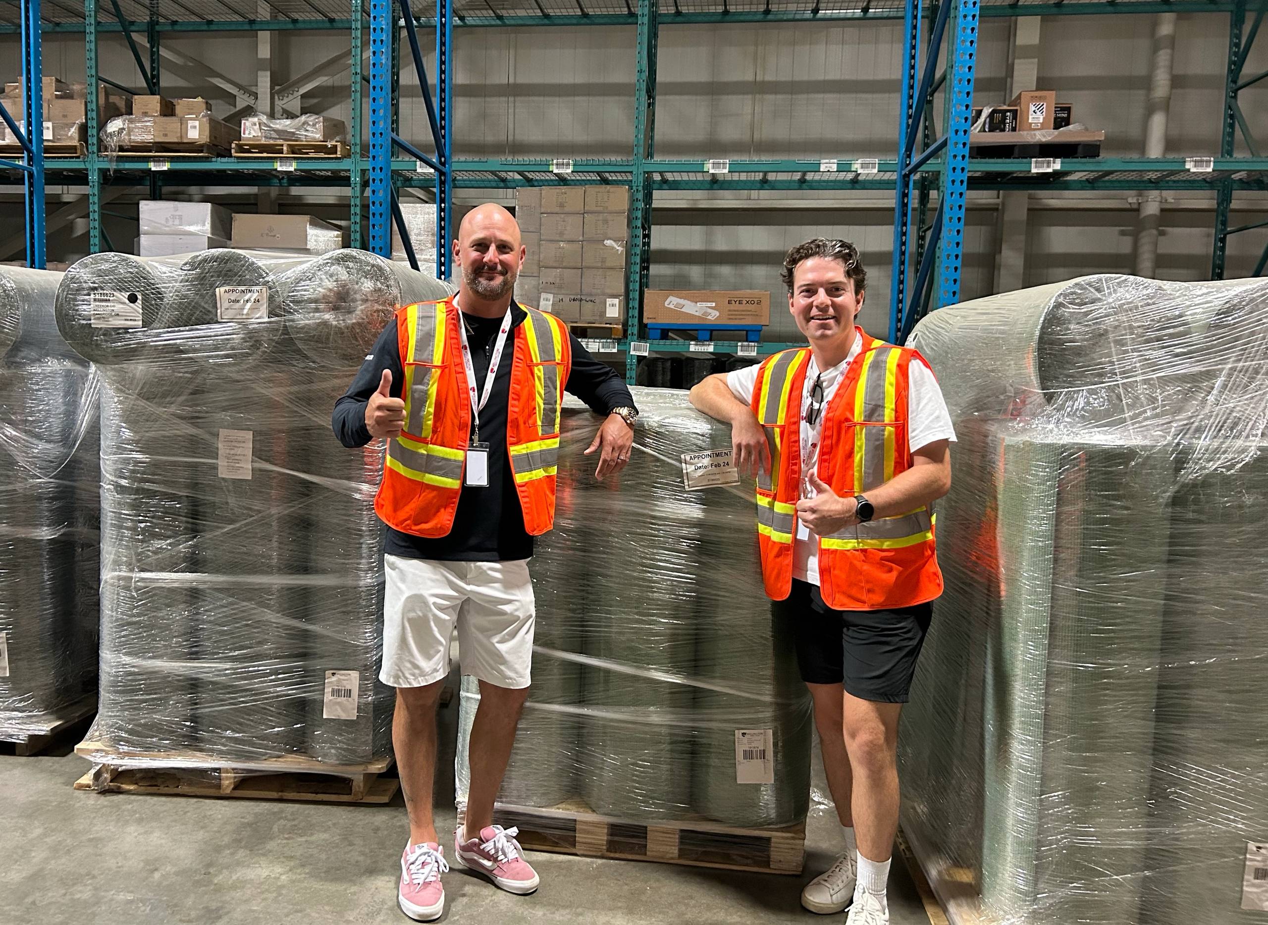 Two men in orange safety vests smile in a warehouse next to pallets of rolled materials.