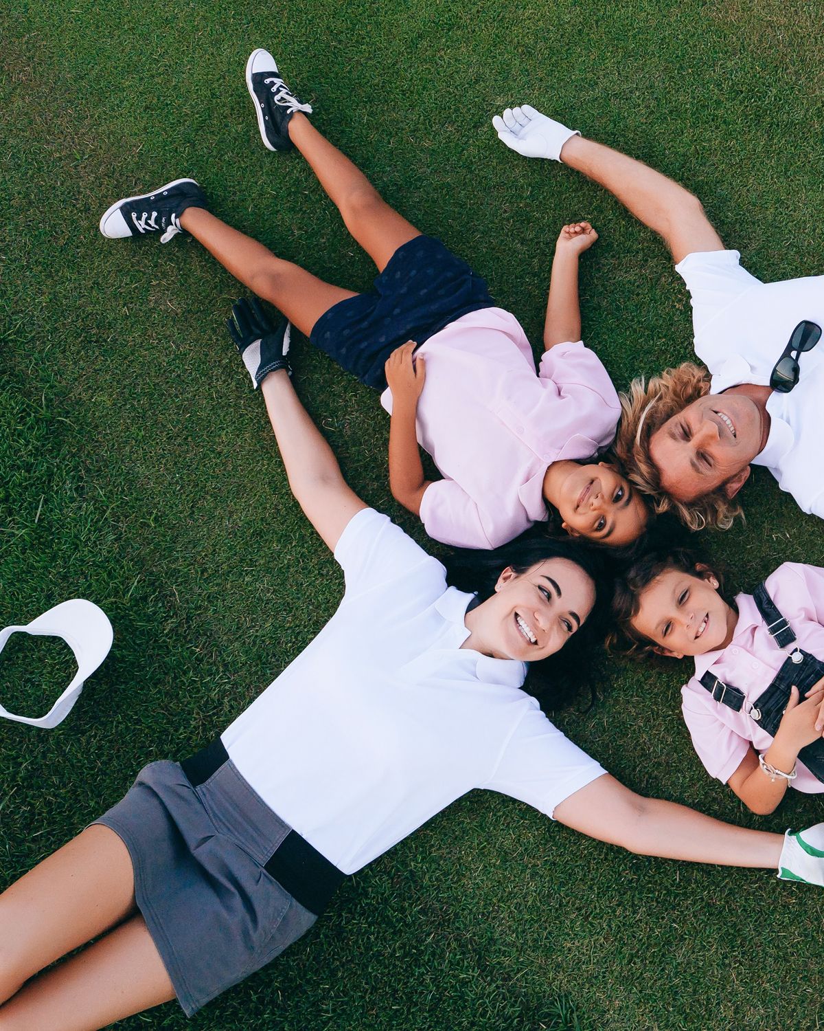 A smiling family of four, in golf attire, lies on green grass with their heads together.