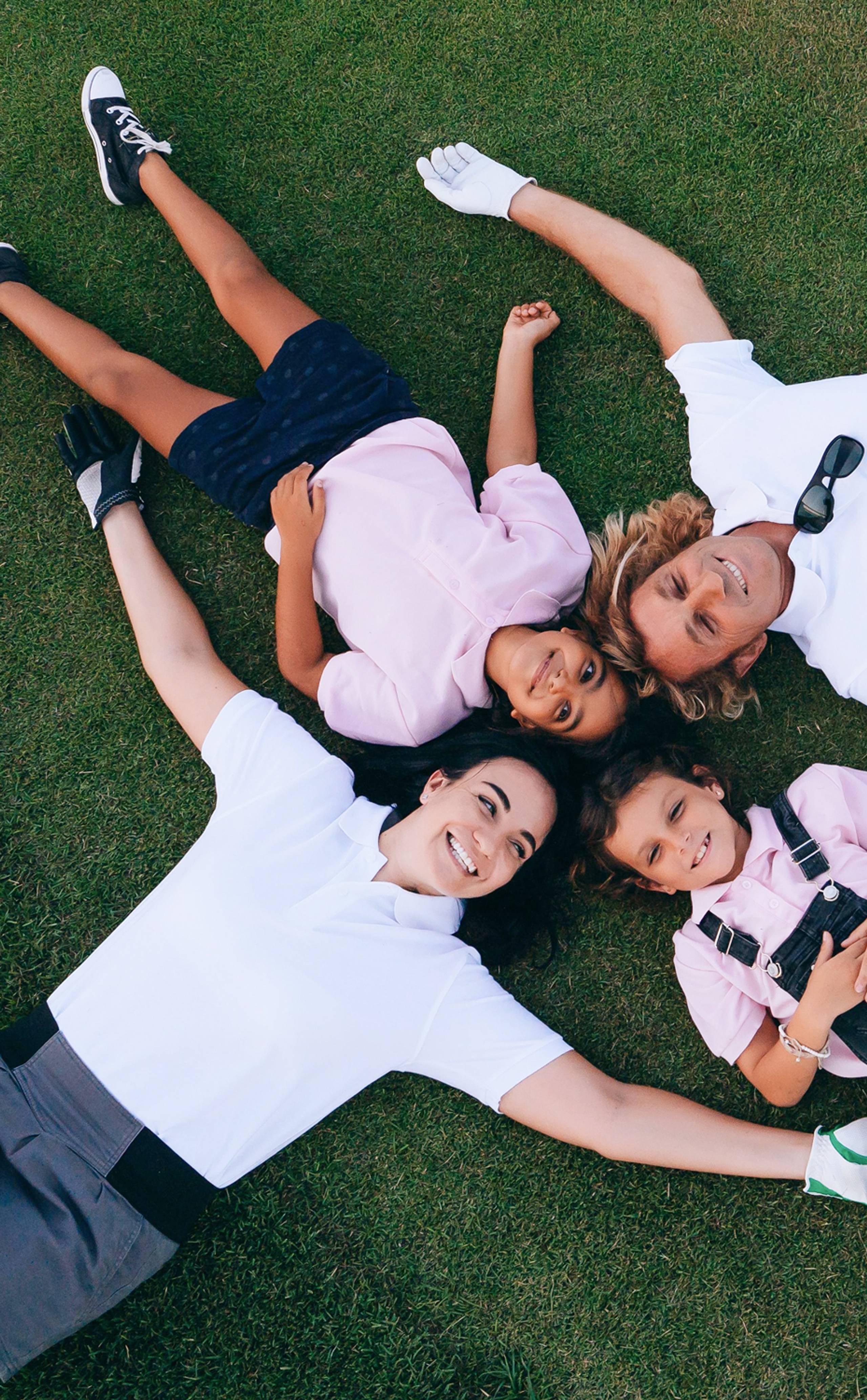 A smiling family of four, in golf attire, lies on green grass with their heads together.