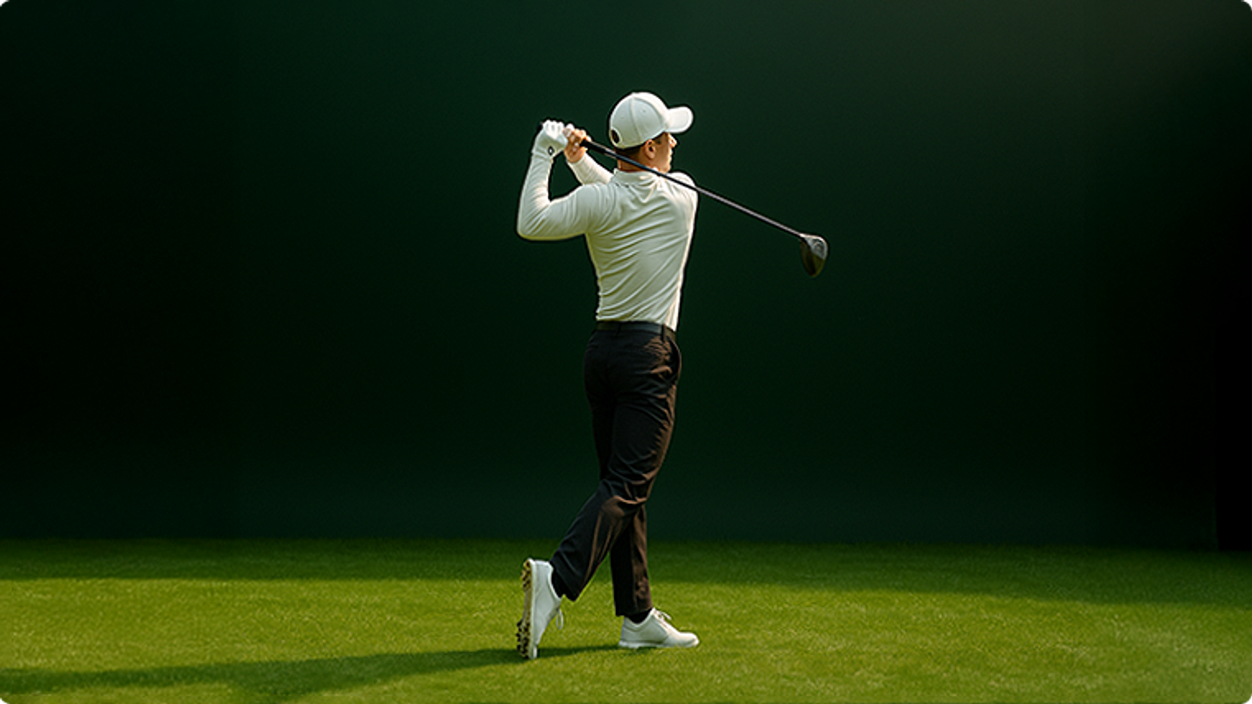 Rear view of a golfer in a white cap and light shirt swinging a driver on a green course.