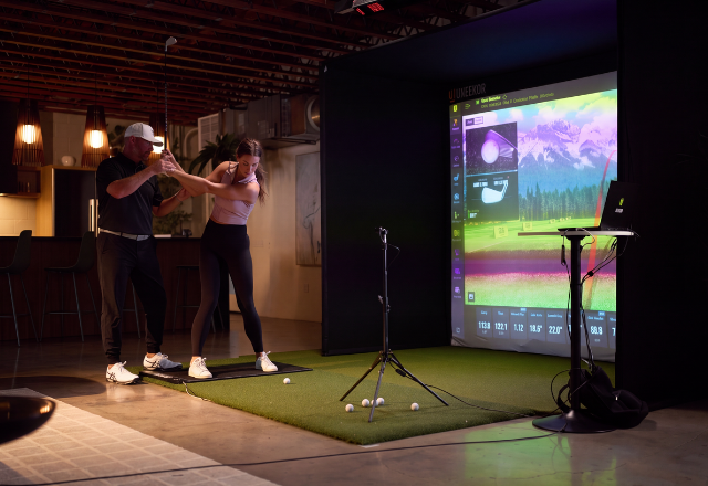 Man coaching woman on golf swing in indoor golf simulator.