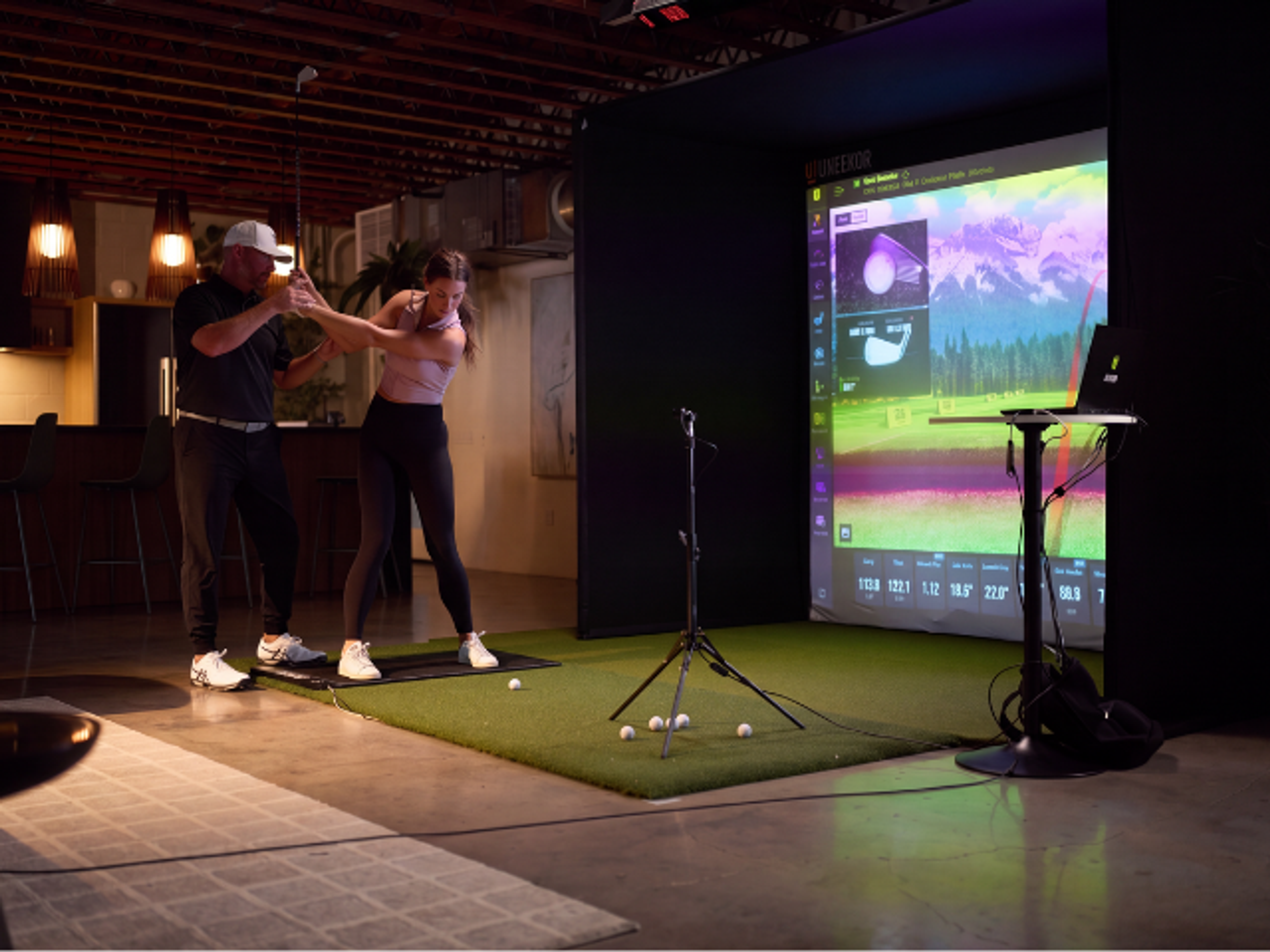 A golf instructor helps a woman with her swing in an indoor golf simulator.