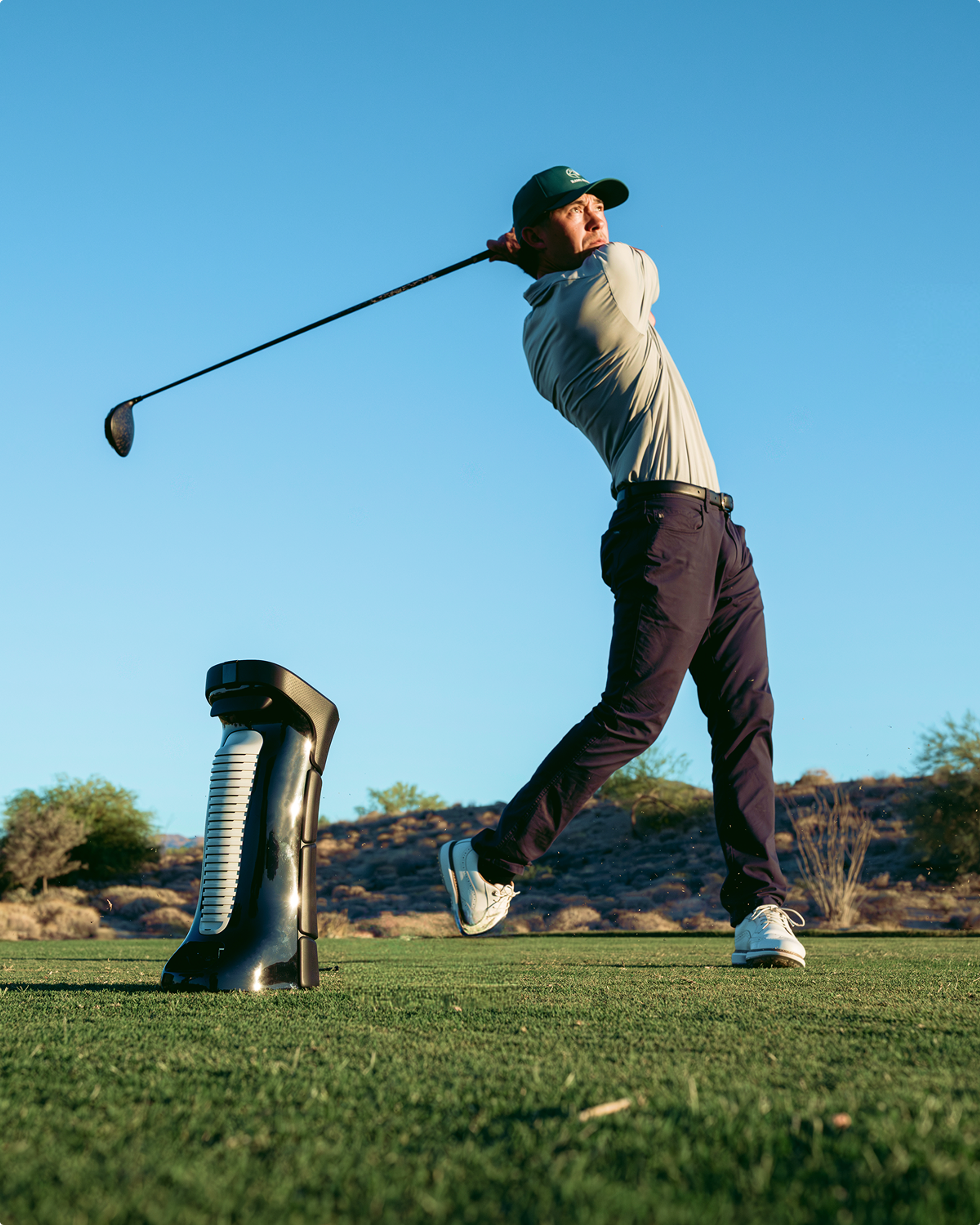 A golfer swings a driver on a sunny course, with a black training device on the grass.