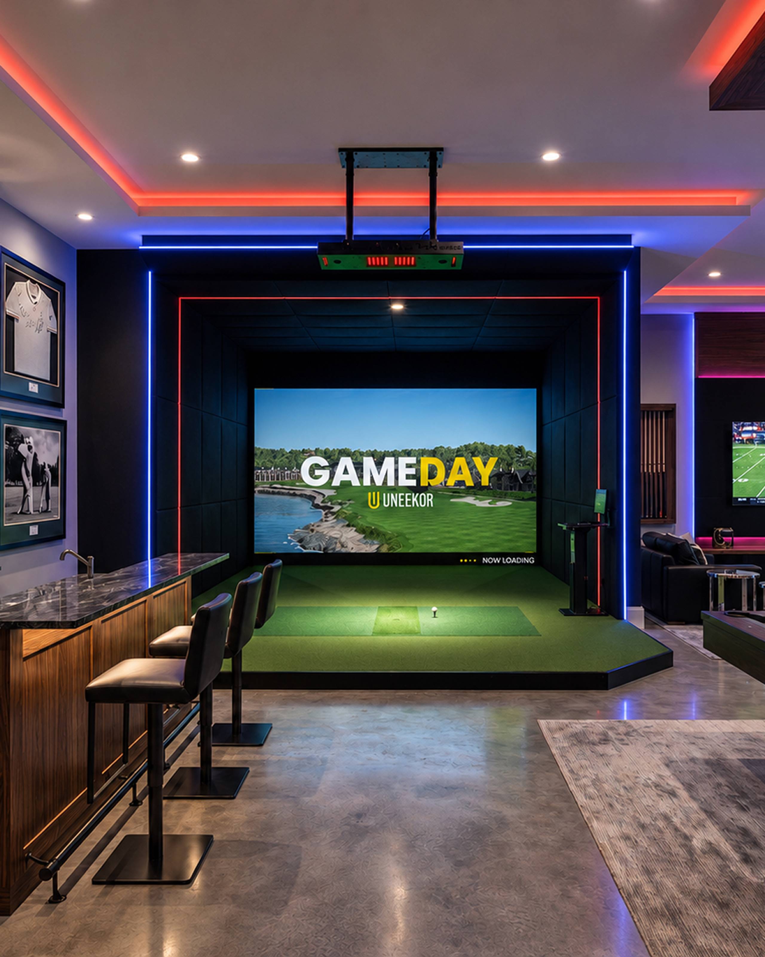 A modern indoor golf simulator room with a large screen, a bar, and colorful LED lighting.