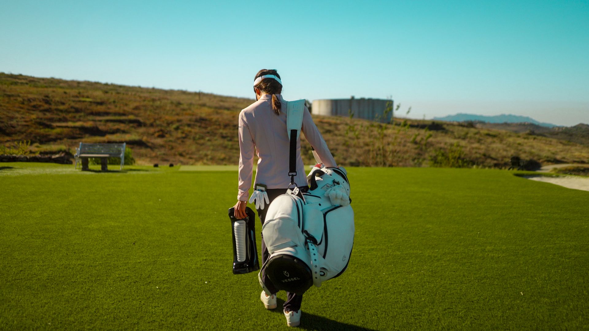 A woman walks across a golf course with a Uneekor EYE MIN Launch Monitor, carrying a white golf bag over her shoulder and holding a water bottle.