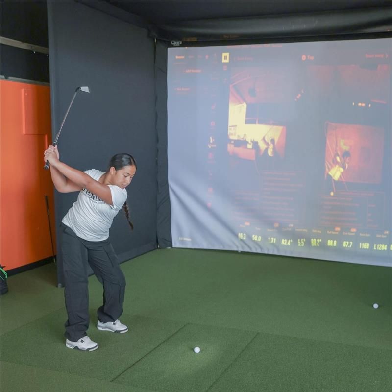 Eine Frau mitten im Golfschwung in einem Golfsimulator mit einem großen Bildschirm, auf dem Schwungdaten angezeigt werden.