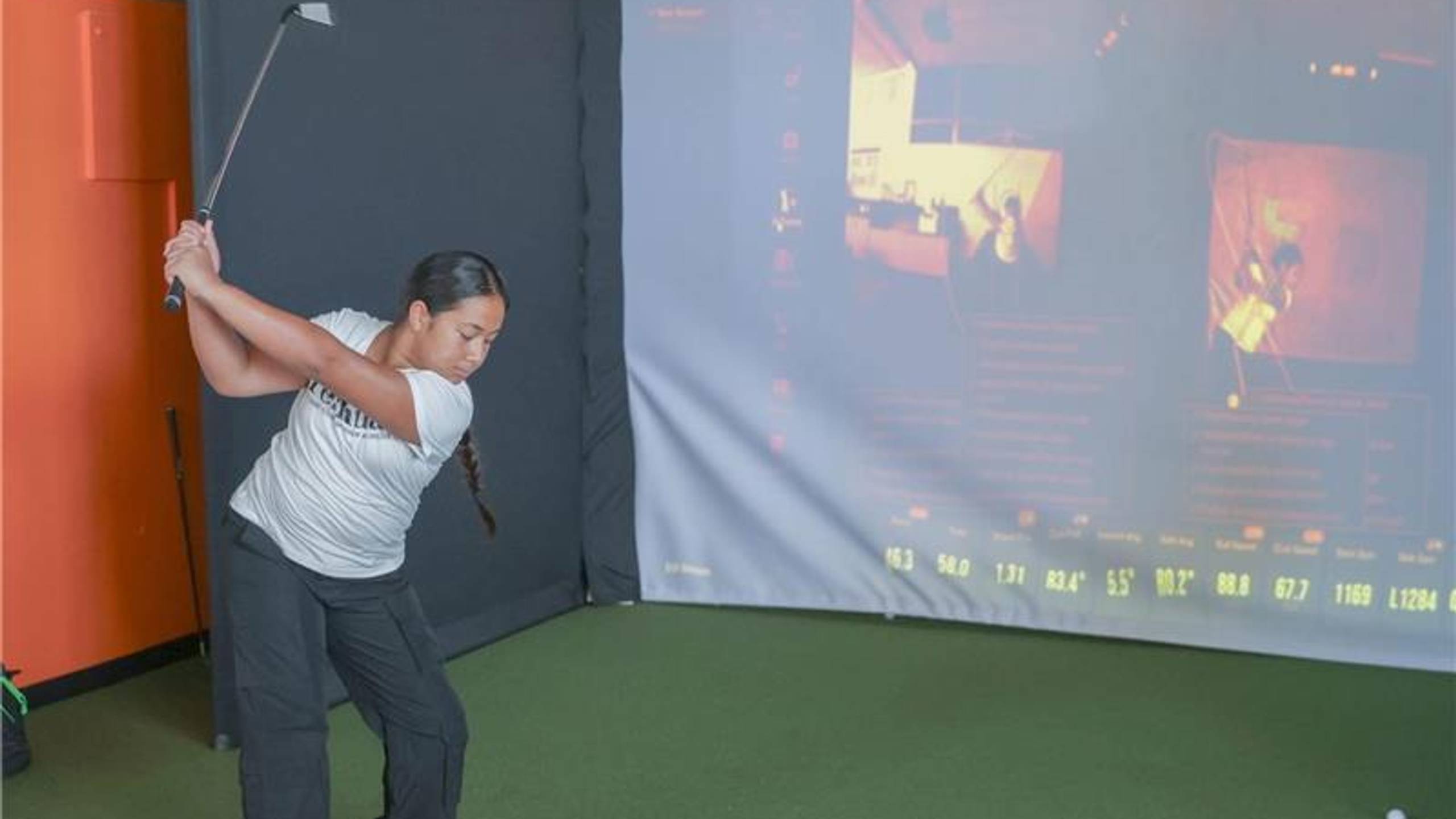 Eine Frau mitten im Golfschwung in einem Golfsimulator mit einem großen Bildschirm, auf dem Schwungdaten angezeigt werden.