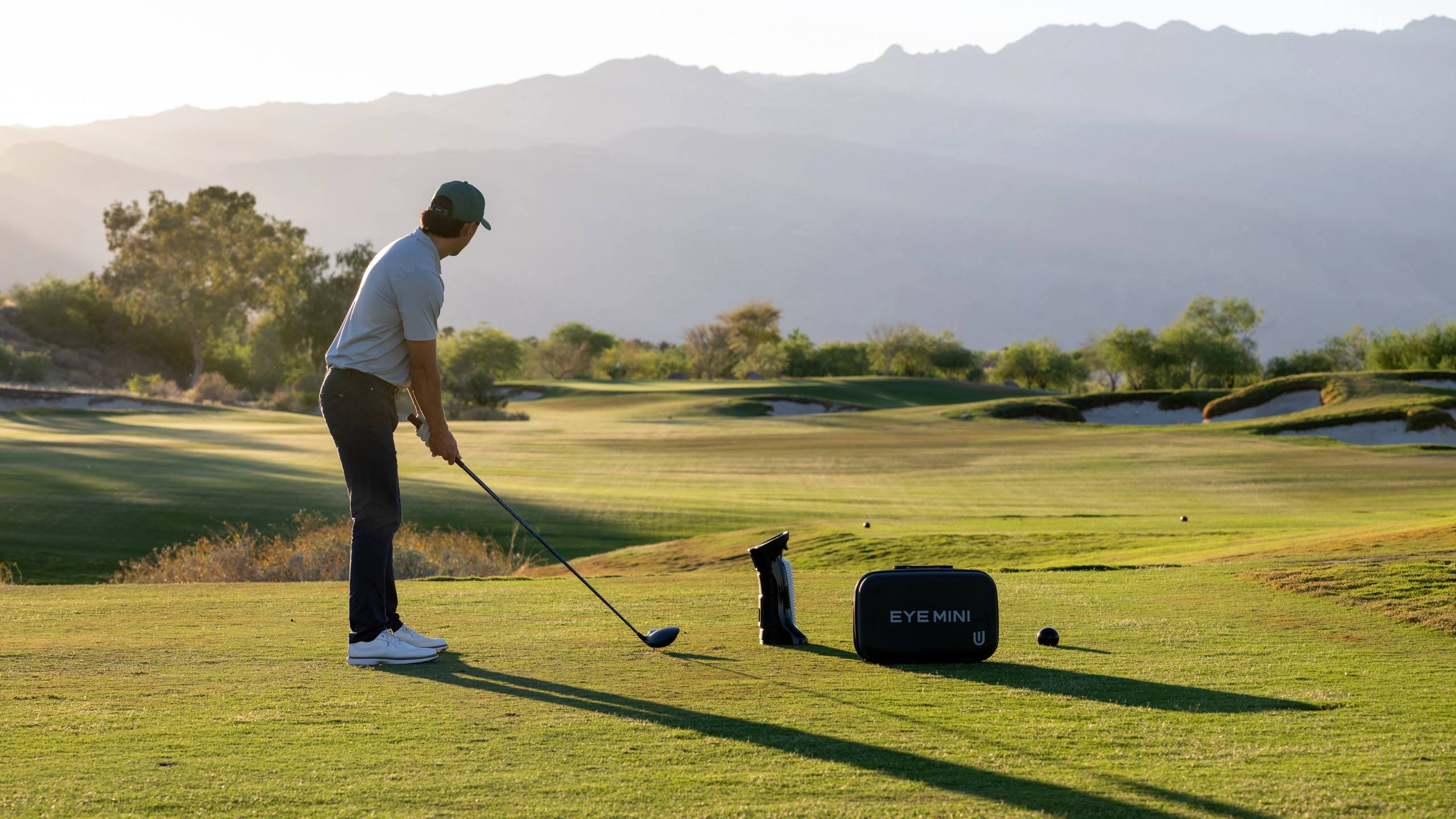 A golfer prepares to swing on a sunny course with mountains in the background, an "EYE MINI" launch monitor beside him.