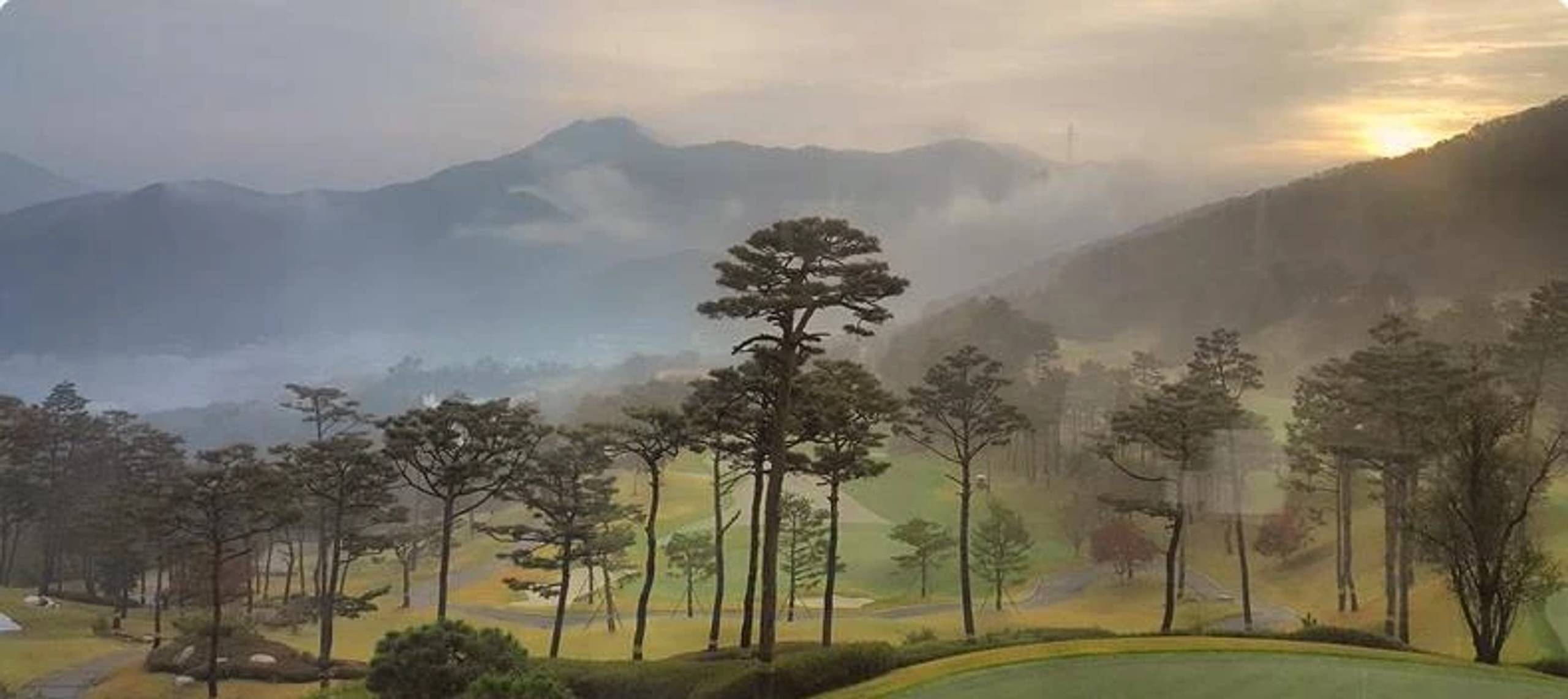 Misty golf course with trees and mountains at sunrise or sunset.
