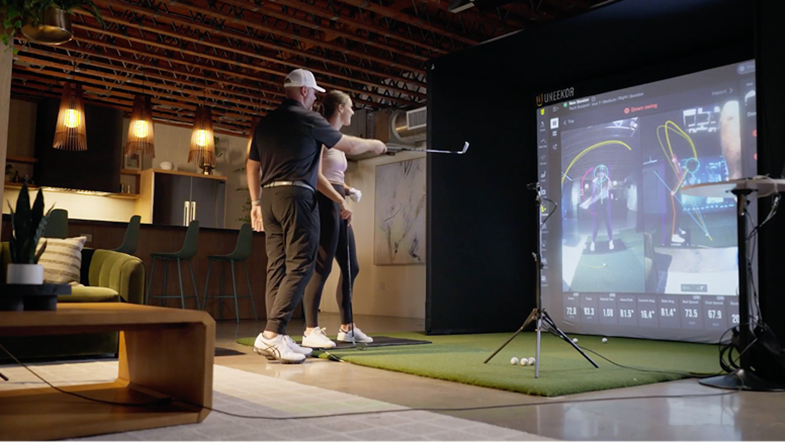 A man coaches a woman on her golf swing in an indoor golf simulator with a large screen displaying swing analytics.
