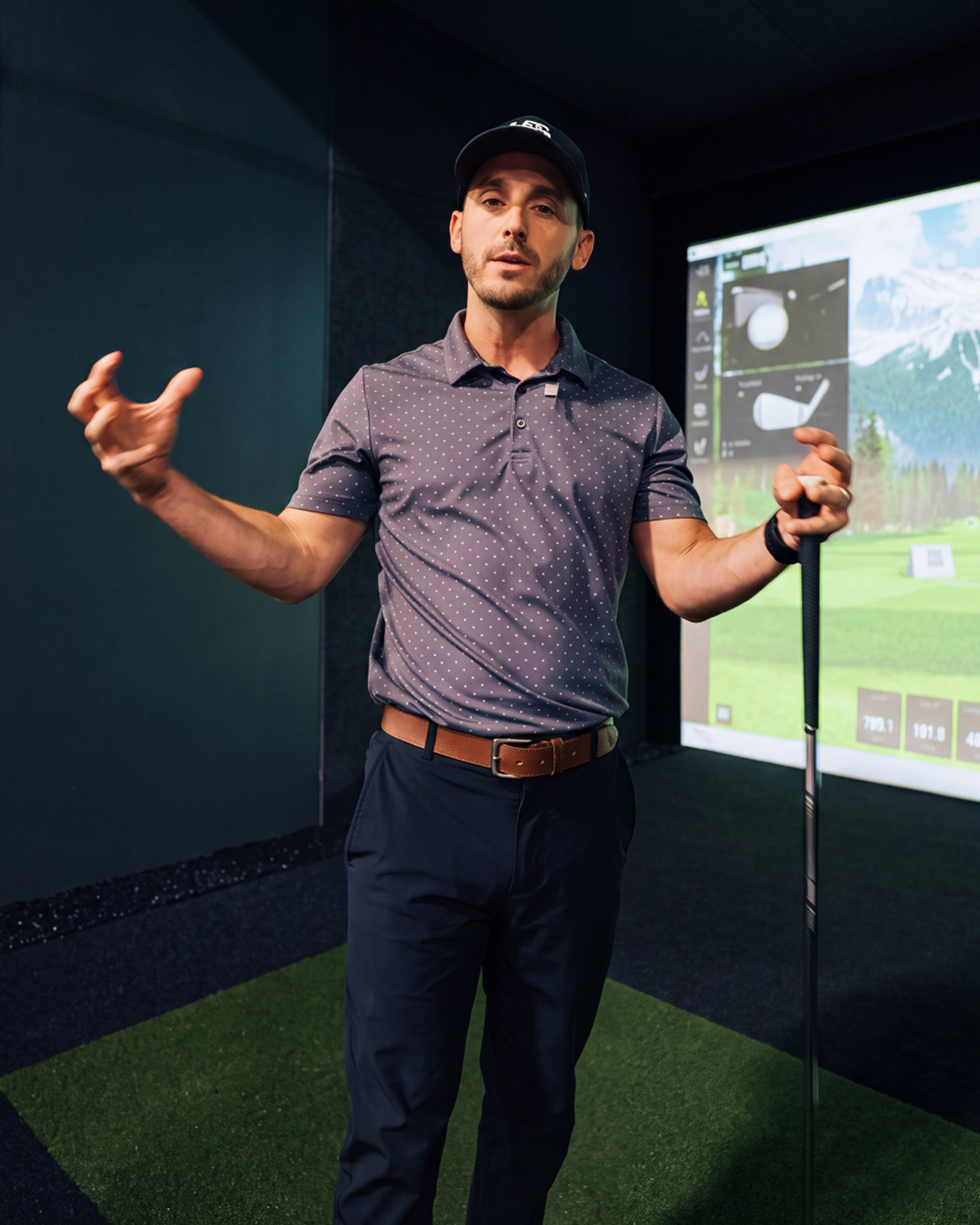 A man holding a golf club gestures in a golf simulator room with a virtual course on screen.