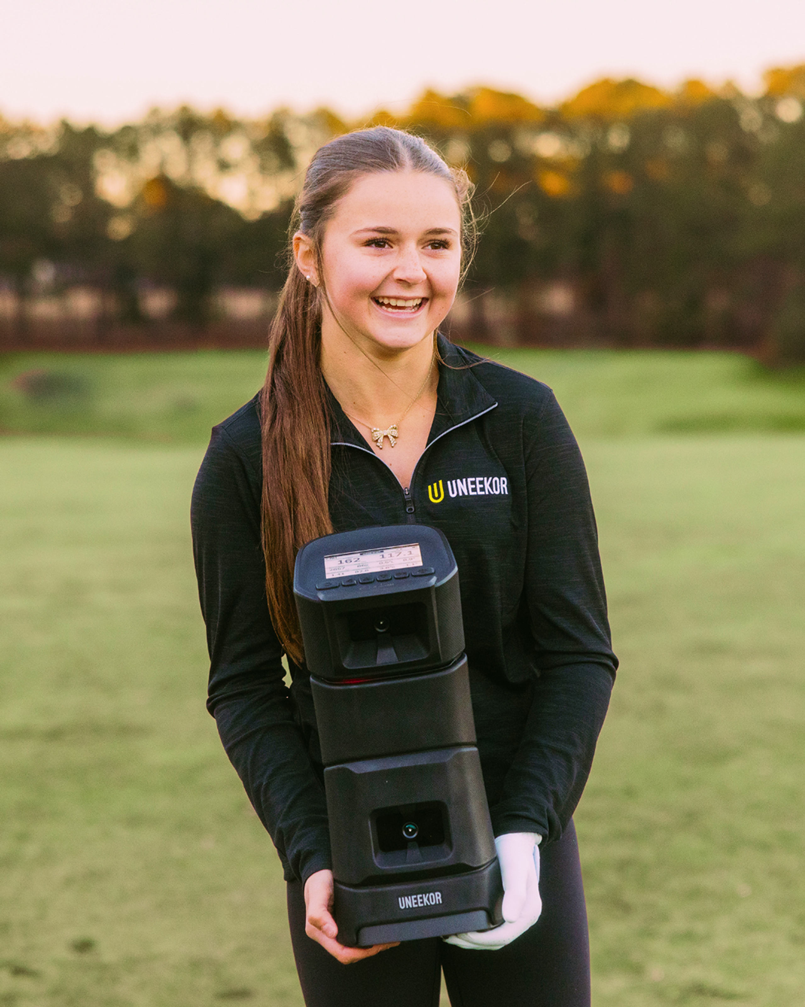 A smiling young woman in a black Uneekor shirt holds a black Uneekor golf launch monitor on a grassy field.