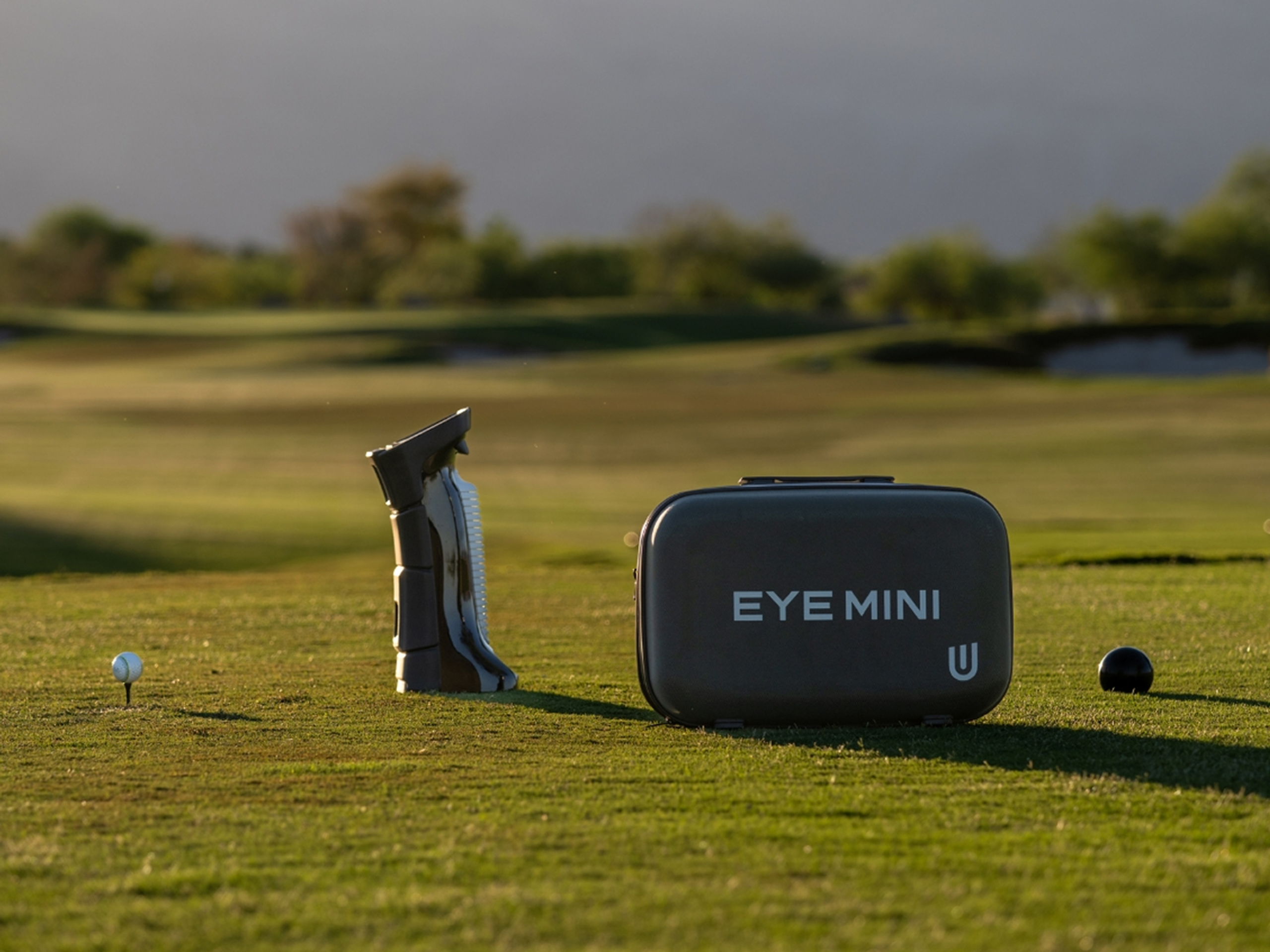 A golf ball on a tee, a golf club cleaner, and an "EYE MINI" device on a golf course.