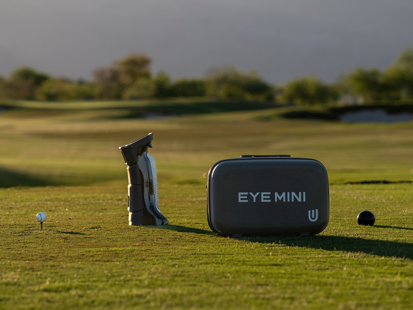 A golf ball on a tee, a golf club cleaner, and an "EYE MINI" device on a golf course.