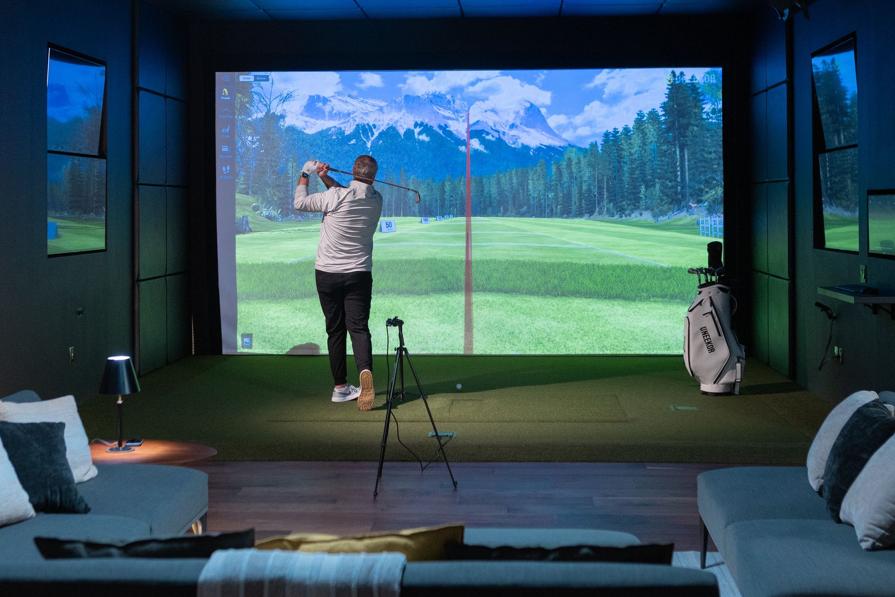 A man golfs in an indoor simulator with a large screen displaying a virtual course and lounge seating.