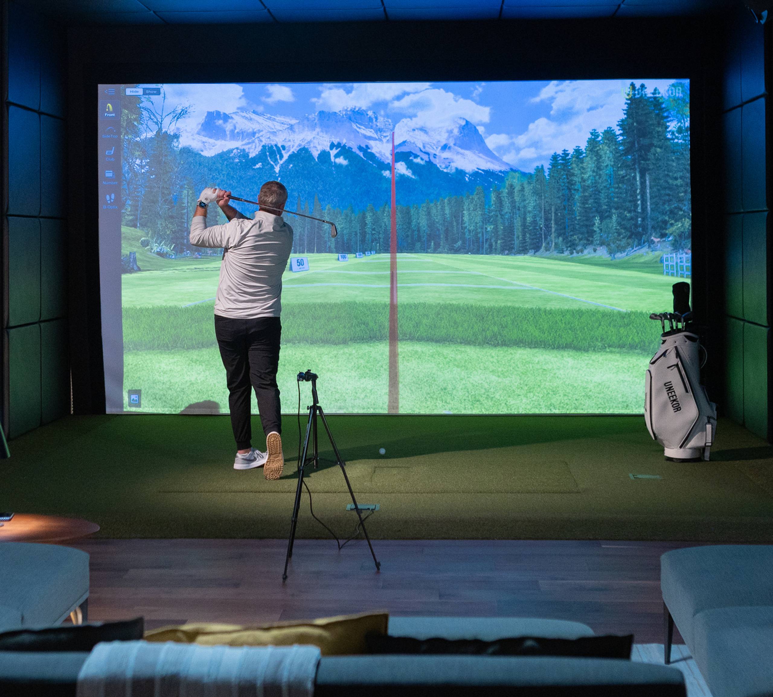 A man golfs in an indoor simulator with a large screen displaying a virtual course and lounge seating.