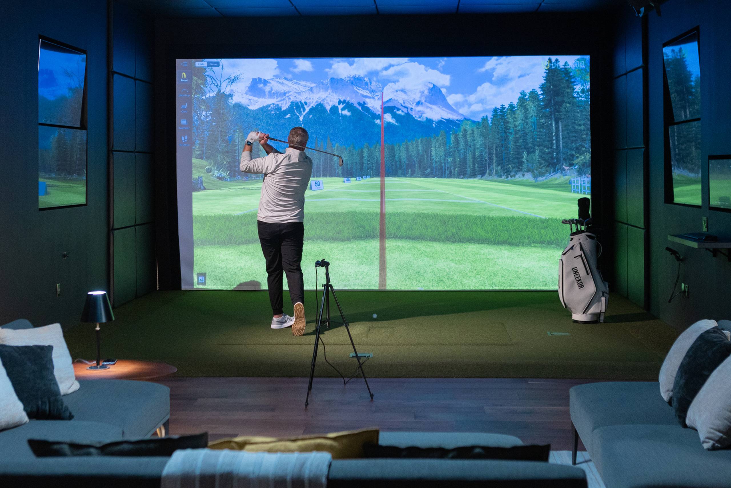 A man swings a golf club in an indoor golf simulator with lounge seating.