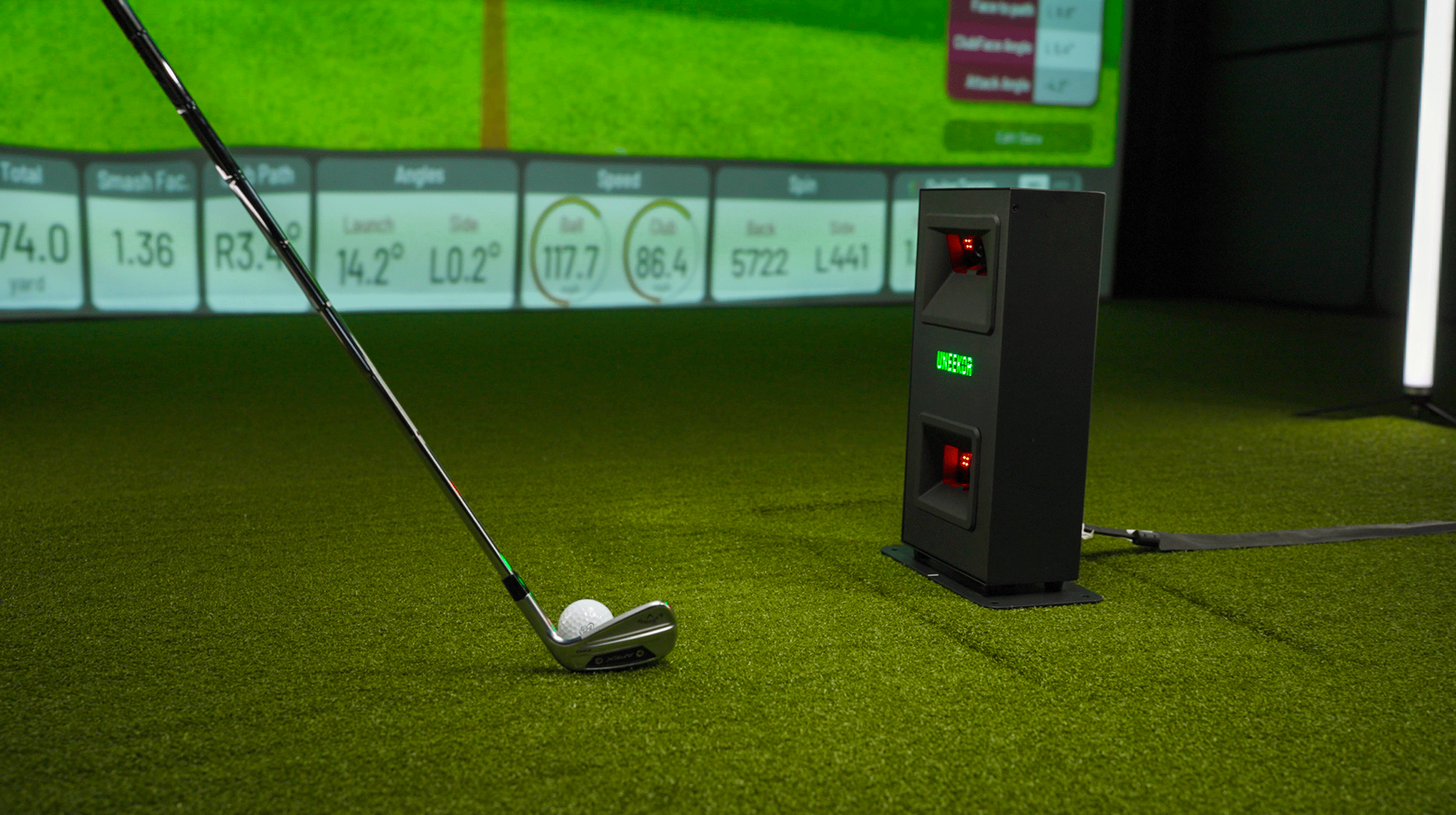 A golf club poised over a ball on artificial turf, with a launch monitor and data display screen in the background.