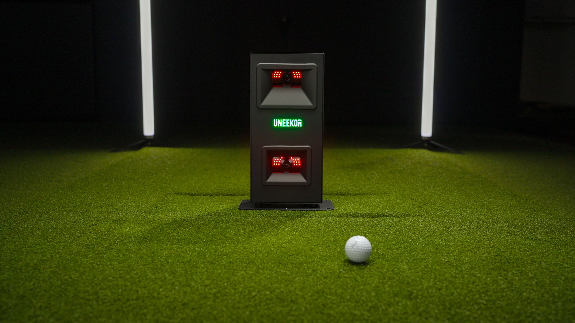 A white golf ball rests on green artificial turf in front of a UNEEKOR golf launch monitor with red lights in an indoor setting.