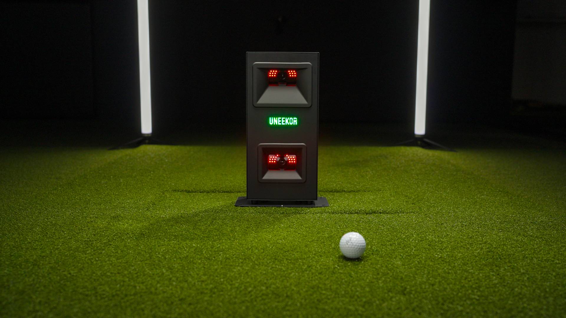 A white golf ball rests on green artificial turf in front of a UNEEKOR golf launch monitor with red lights in an indoor setting.