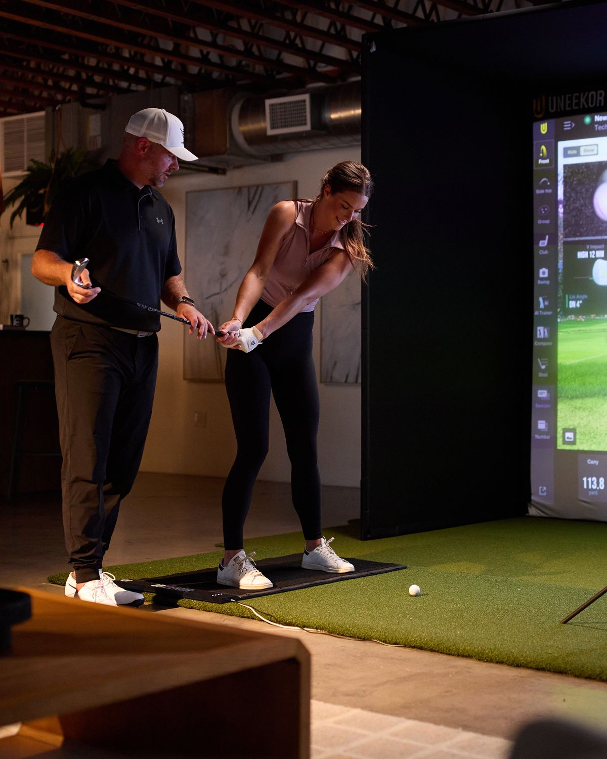 A golf instructor coaches a woman practicing her swing in a golf simulator.