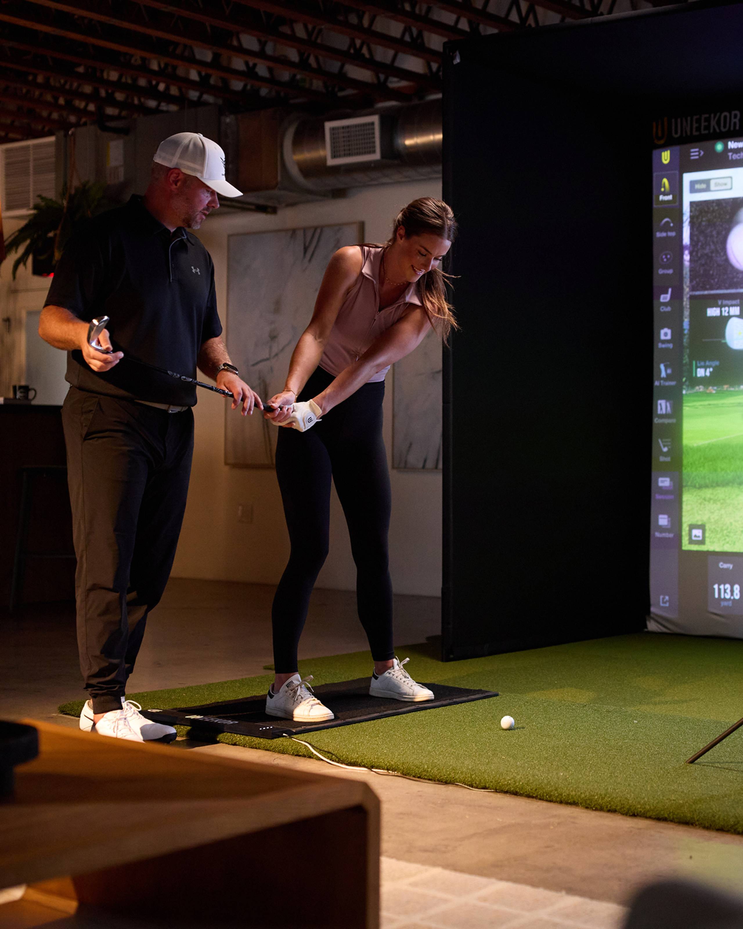 A man coaches a woman's golf swing in an indoor golf simulator.