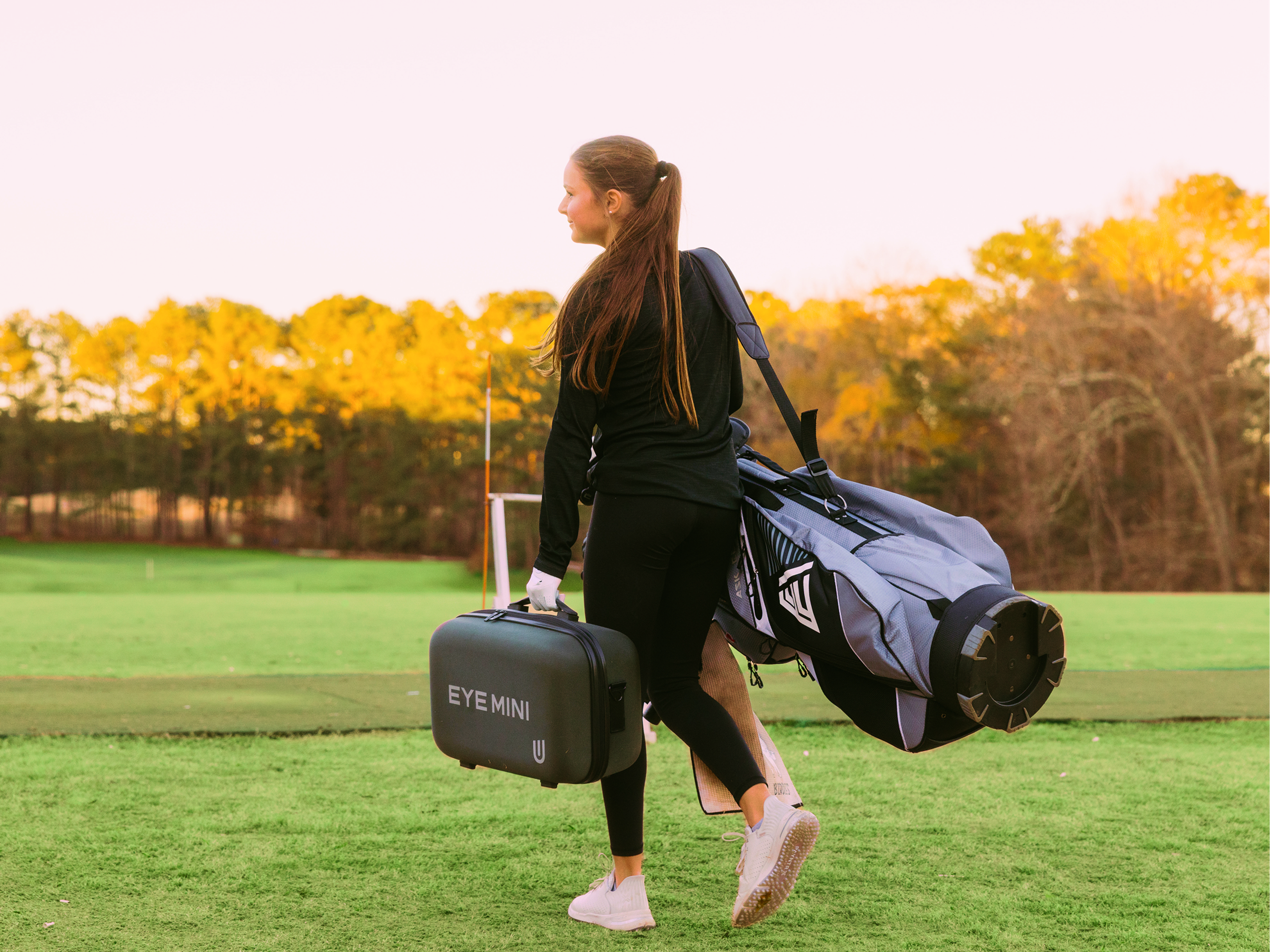 Woman carrying a golf bag and an "EYE MINI" case on a golf course.