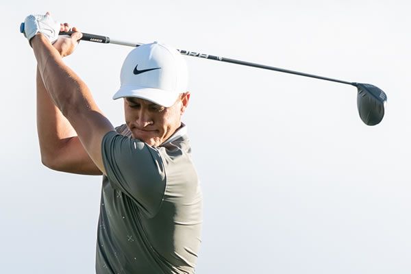 Male golfer in a white Nike cap and grey shirt at the top of his golf swing.