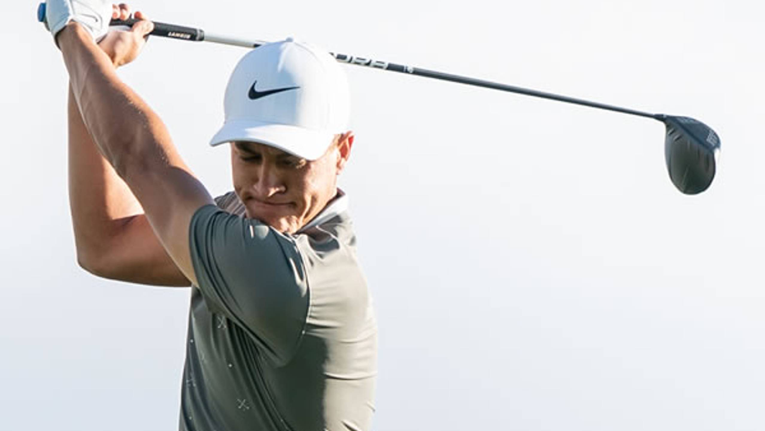 Male golfer in a white Nike cap and grey shirt at the top of his golf swing.